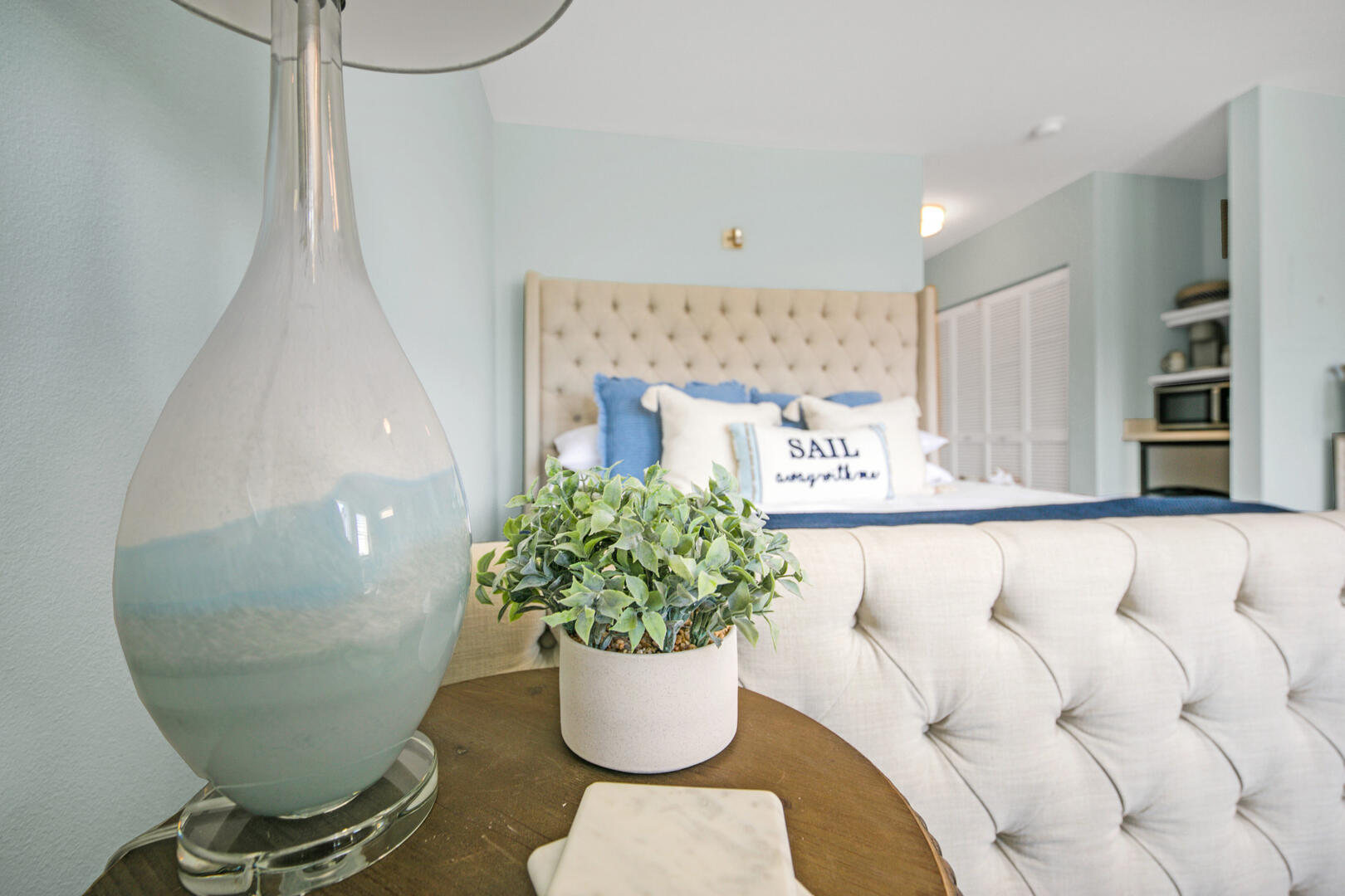 Staged bedroom with a tufted cream bed, blue pillows, and a large glass lamp in the foreground.