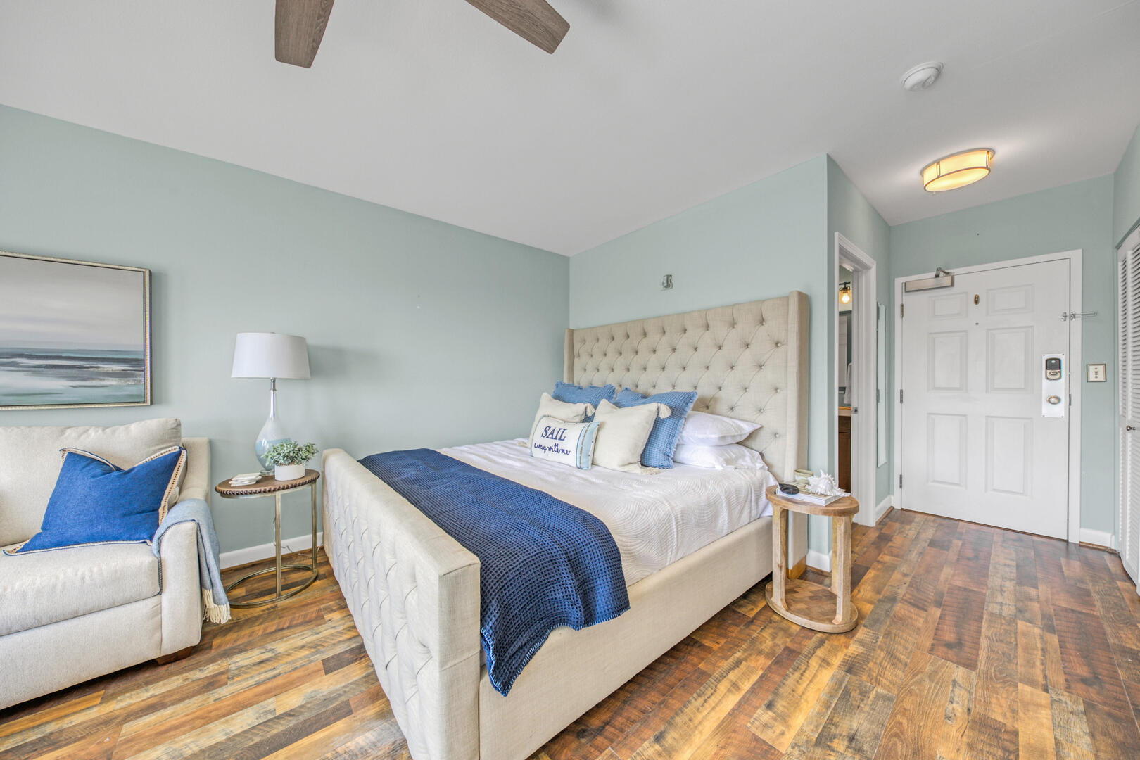 Furnished bedroom with a cream tufted bed, blue accents, an armchair, and wood-look flooring.