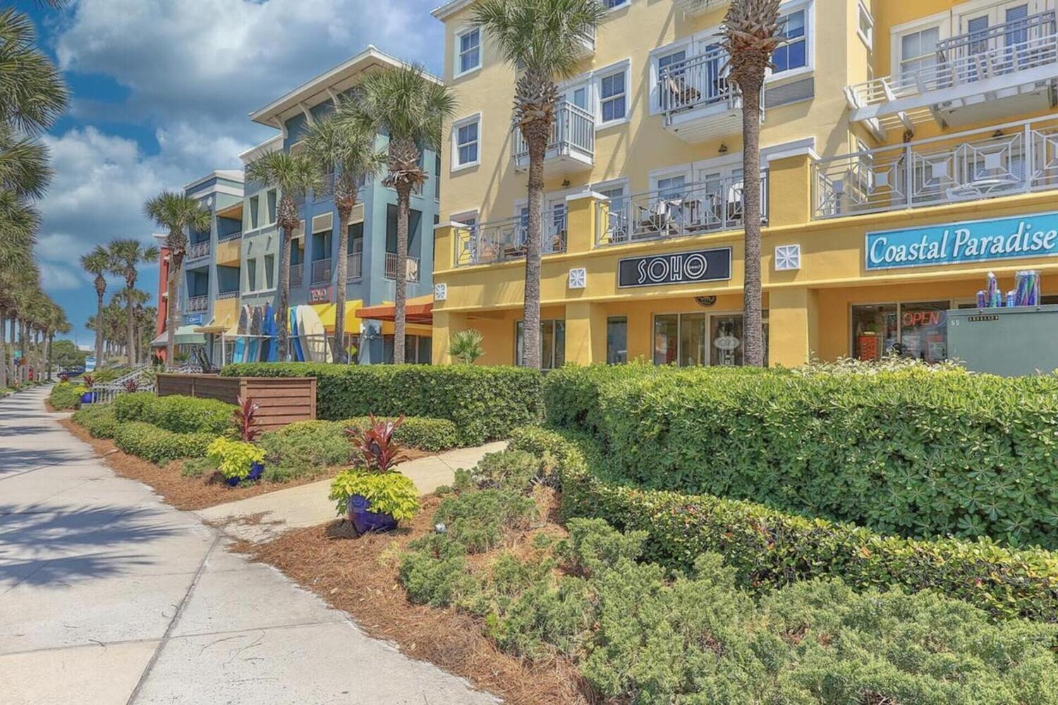 Multi-story yellow building with balconies, palm trees, and ground-floor shops along a landscaped sidewalk.