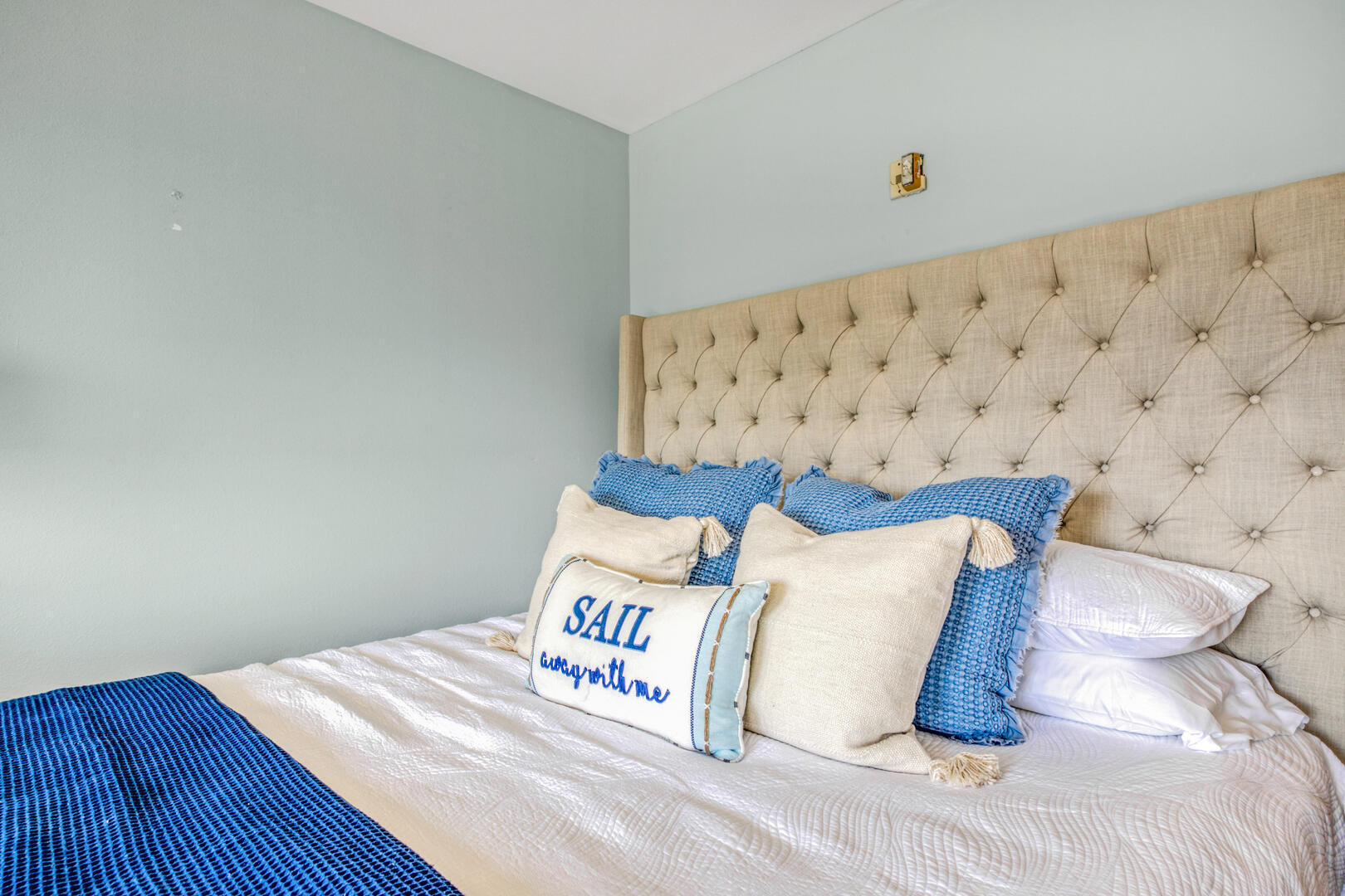 Bedroom with a tall tufted headboard and blue-and-cream pillows, including one embroidered “SAIL away with me.”