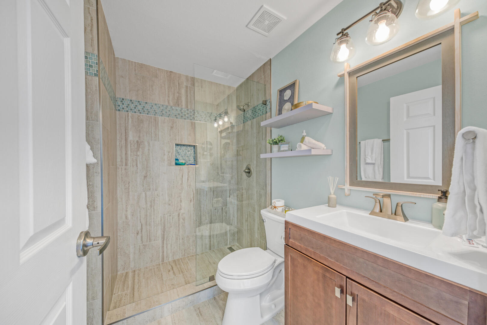 Bathroom with glass-enclosed tiled shower, blue mosaic accents, toilet, and wood vanity.