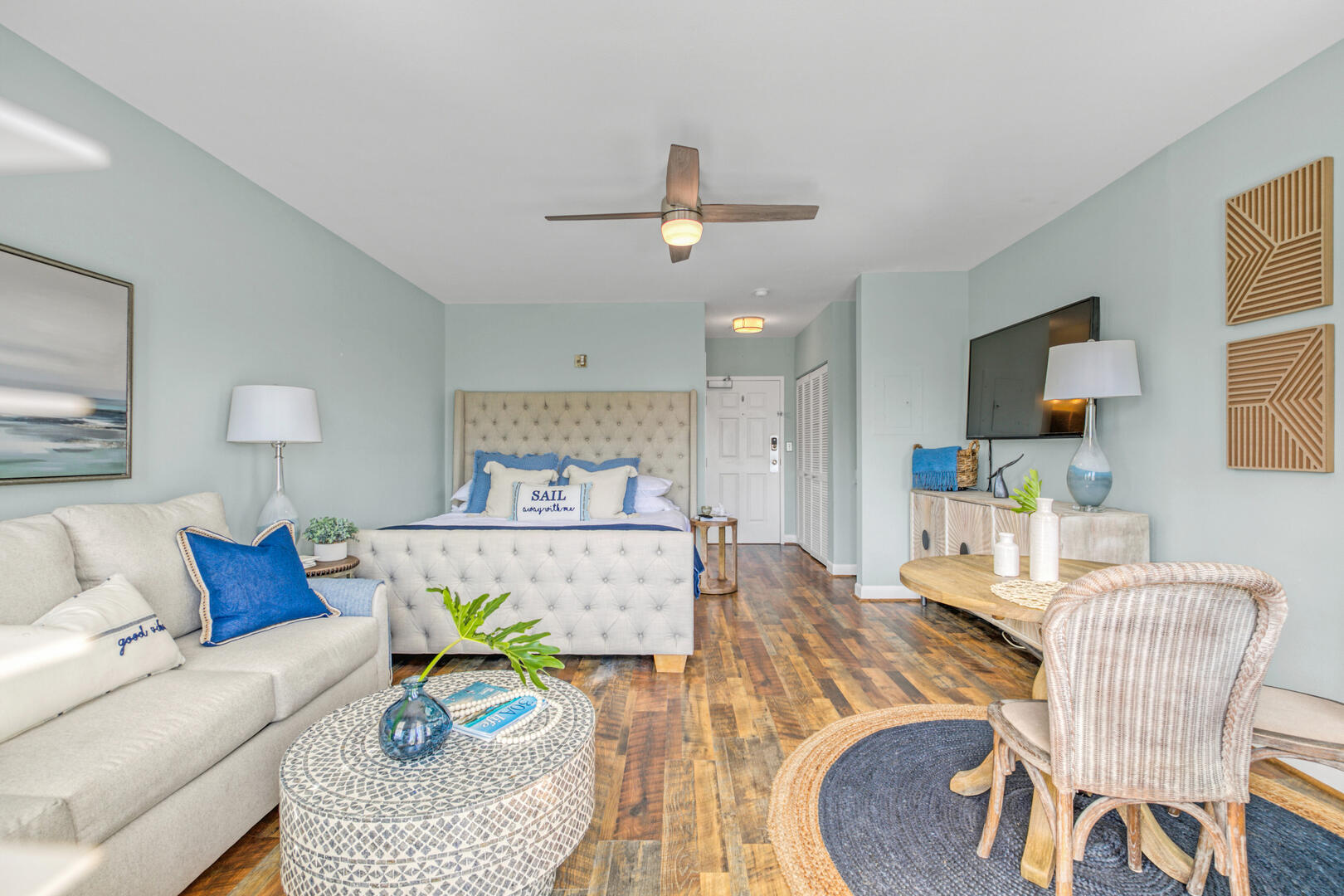 Staged studio bedroom and living area with a tufted bed, cream sofa, and wicker seating.