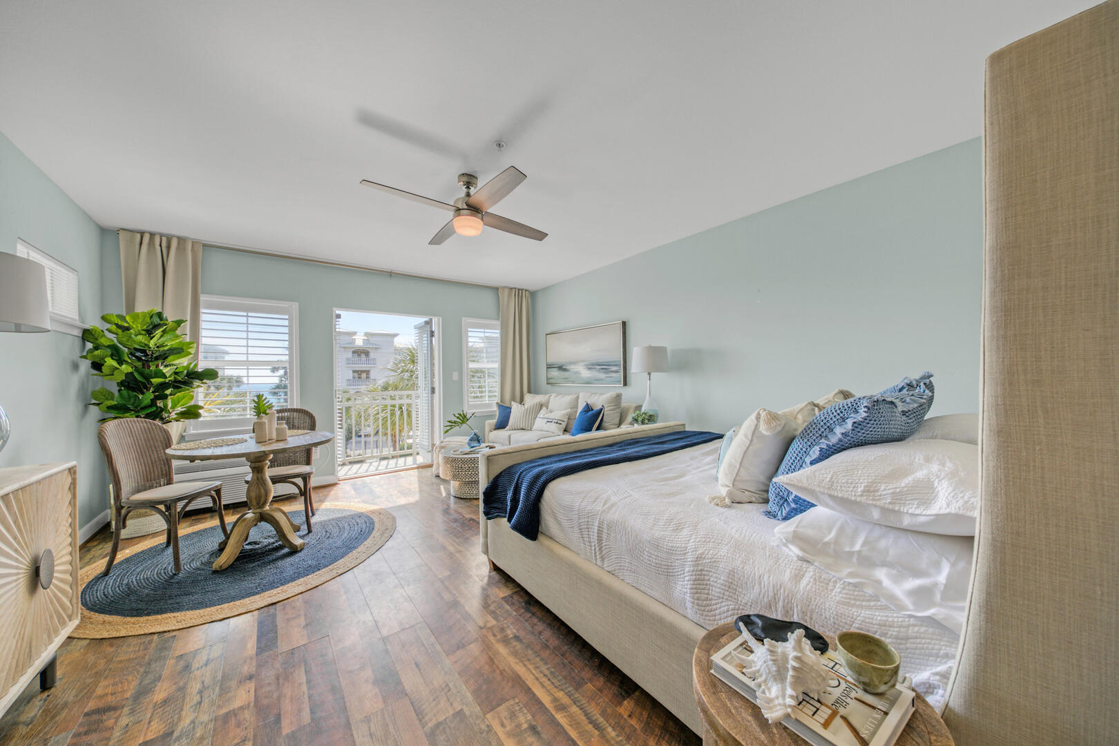 Staged bedroom with a white bed, blue accents, and a balcony door with a glimpse of water.