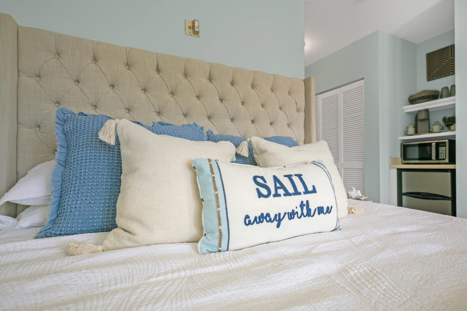 Bedroom with a tall tufted headboard, blue and cream pillows, and a “SAIL away with me” cushion.