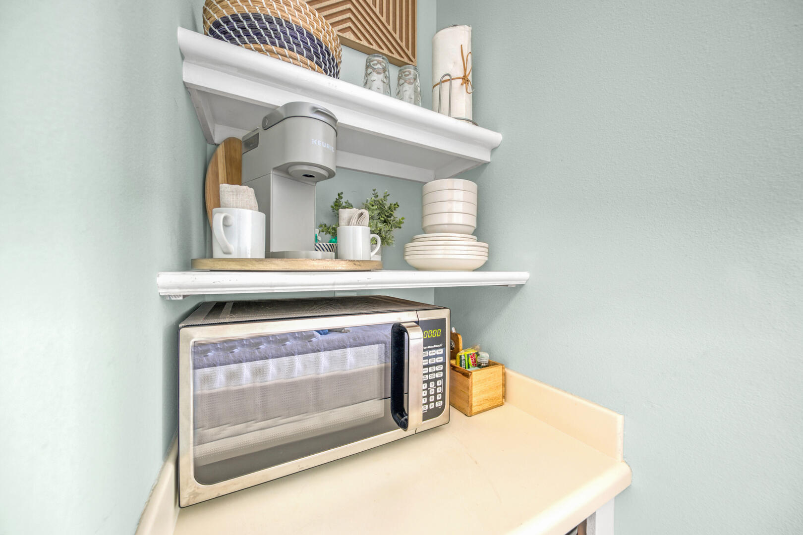 Kitchen counter with a microwave beneath shelves holding a coffee maker, mugs, and stacked dishes.