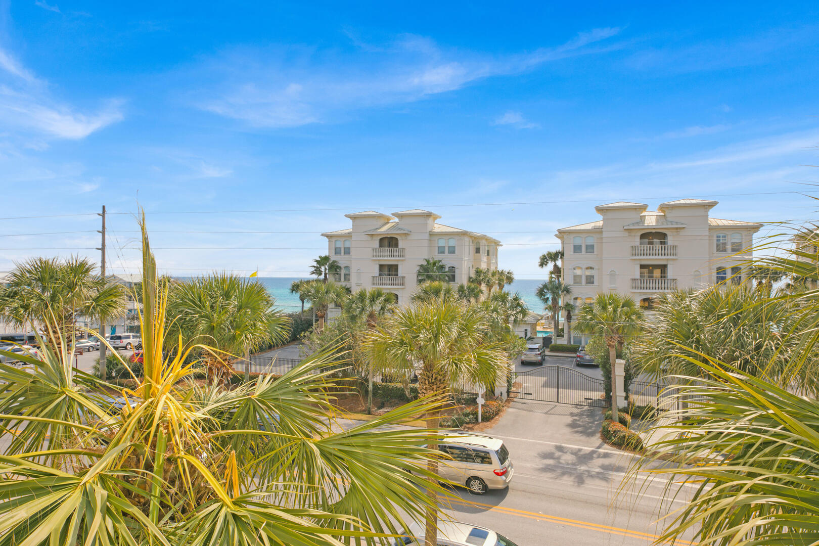 White condominium buildings and palm trees beside the turquoise Gulf, viewed across a gated driveway.