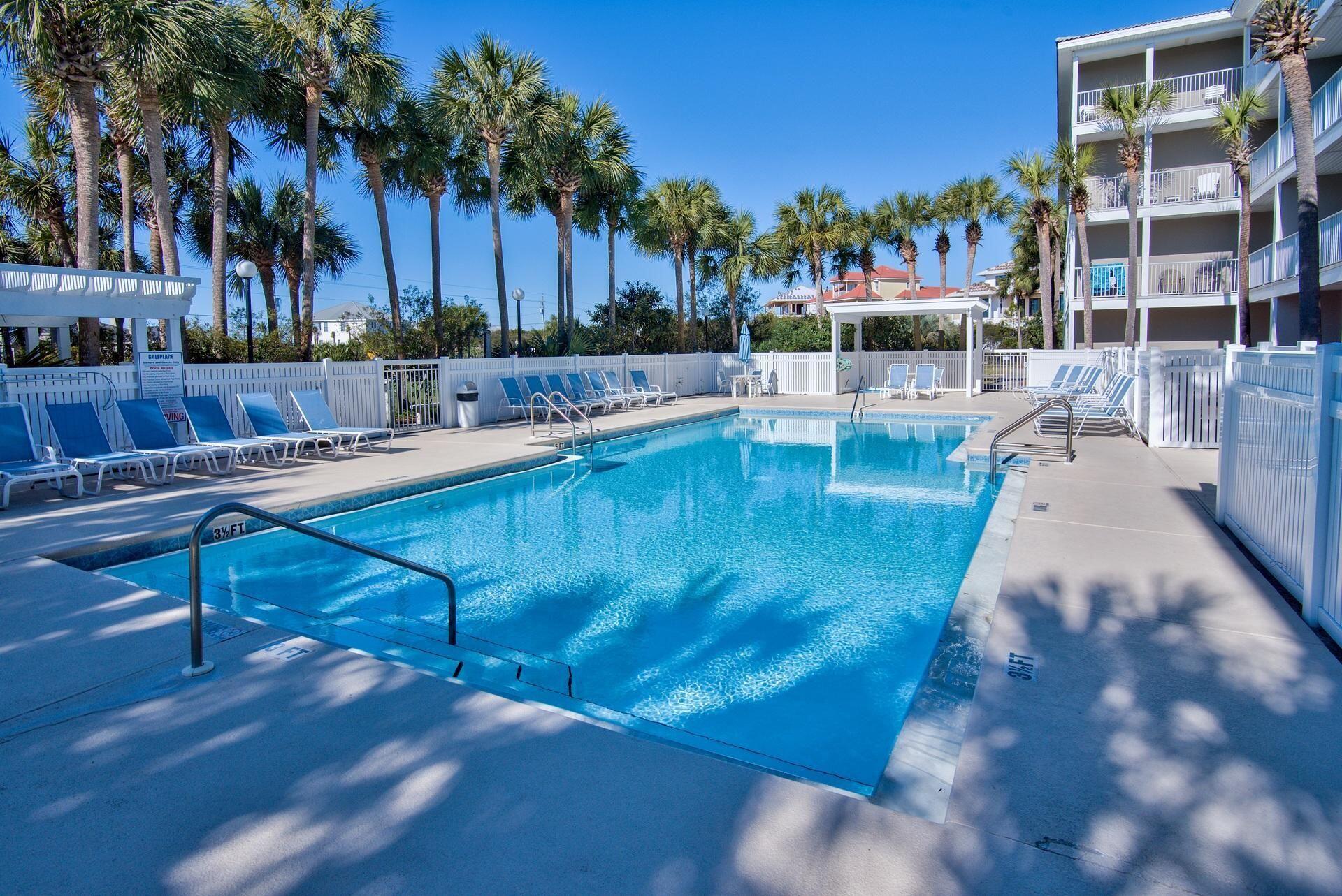 Condominium pool with palm trees, white lounge chairs, and a multistory building with balconies.