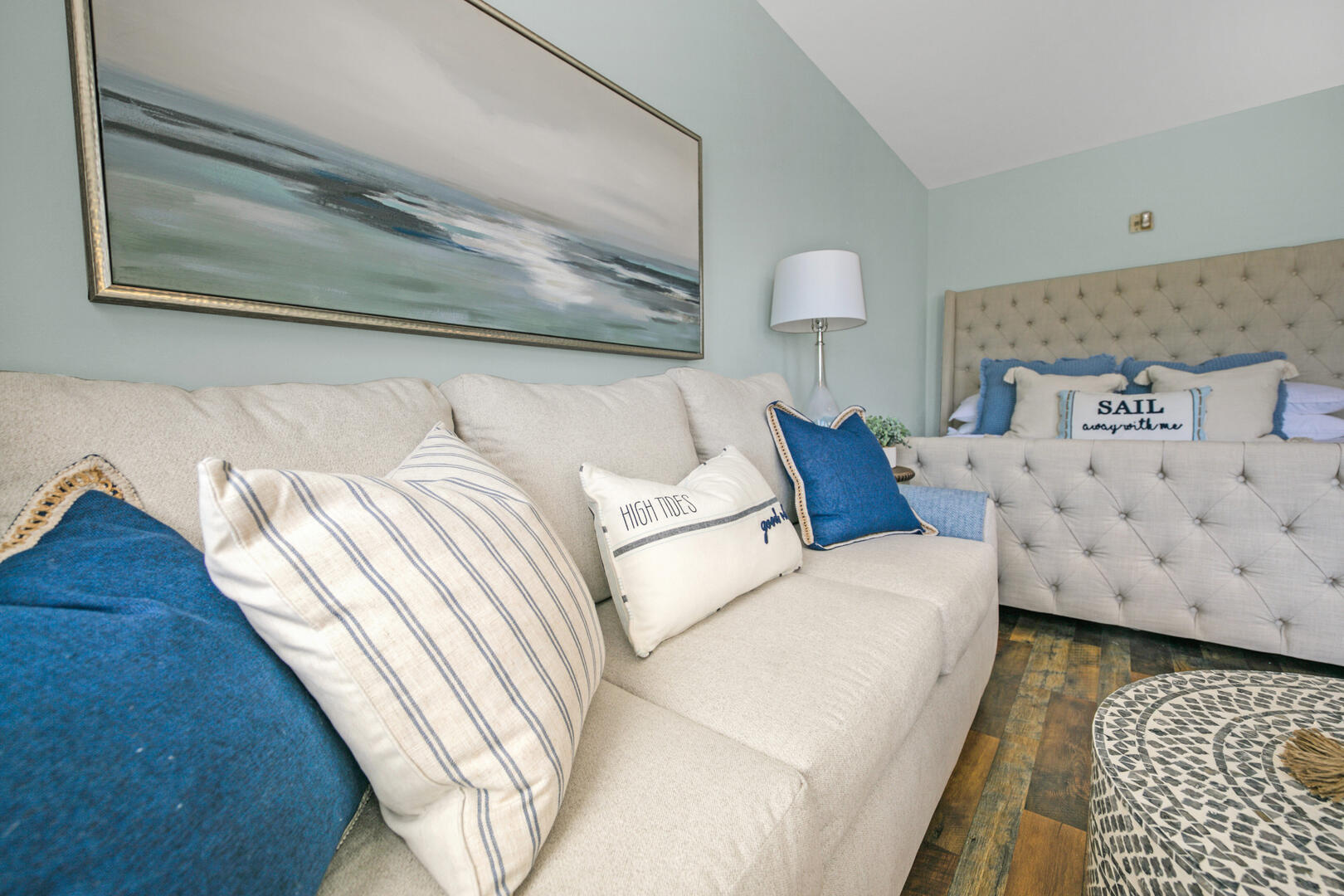 Living room with a cream sofa, blue-and-white pillows, and a bed beneath coastal artwork.