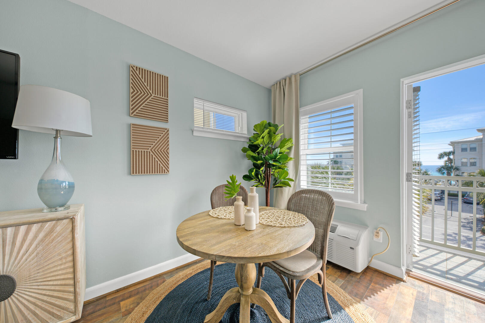Dining nook with a round wood table and balcony doors overlooking a glimpse of ocean.