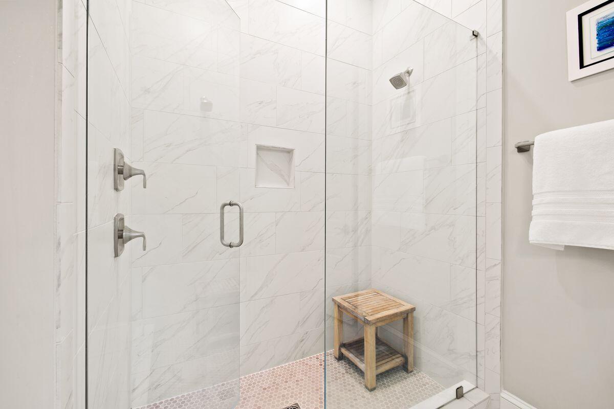 Walk-in shower with white marble-look tile, frameless glass, mosaic flooring, and a wooden stool.