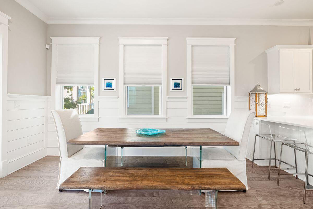 Dining room with a rustic wood table, white chairs, and three tall windows with blinds.