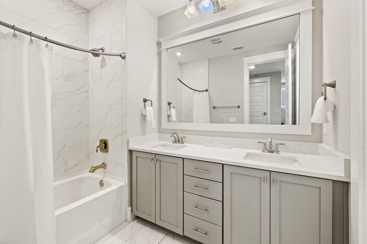 Bathroom with a gray double vanity, large mirror, and marble-look tile around the bathtub.