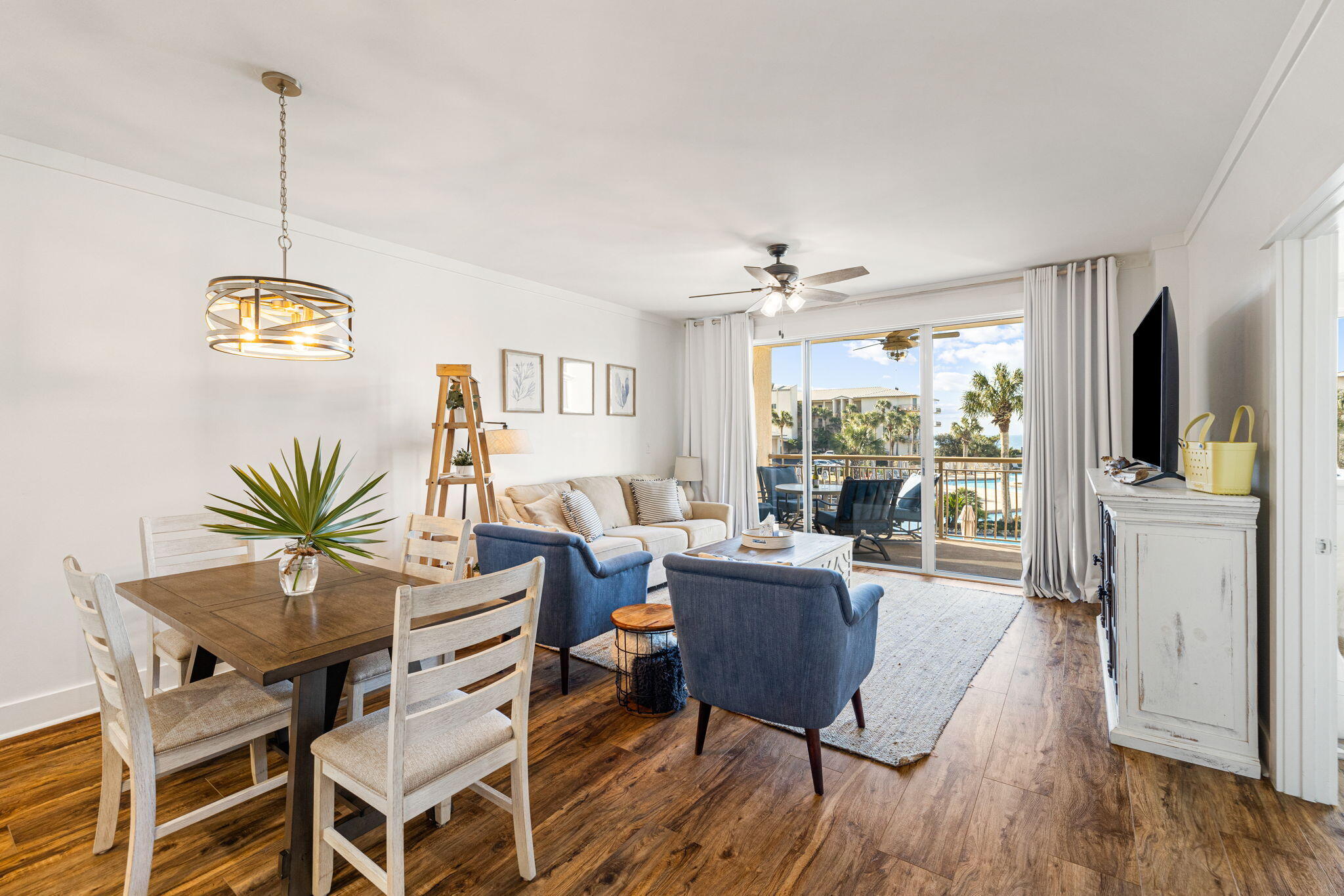 Furnished condo living and dining area opening onto a balcony with palm trees and a pool view.