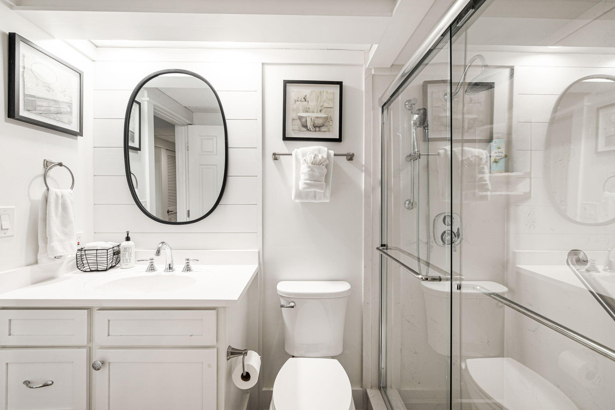 Bright white bathroom with a glass shower, vanity, oval mirror, and horizontal wall paneling.