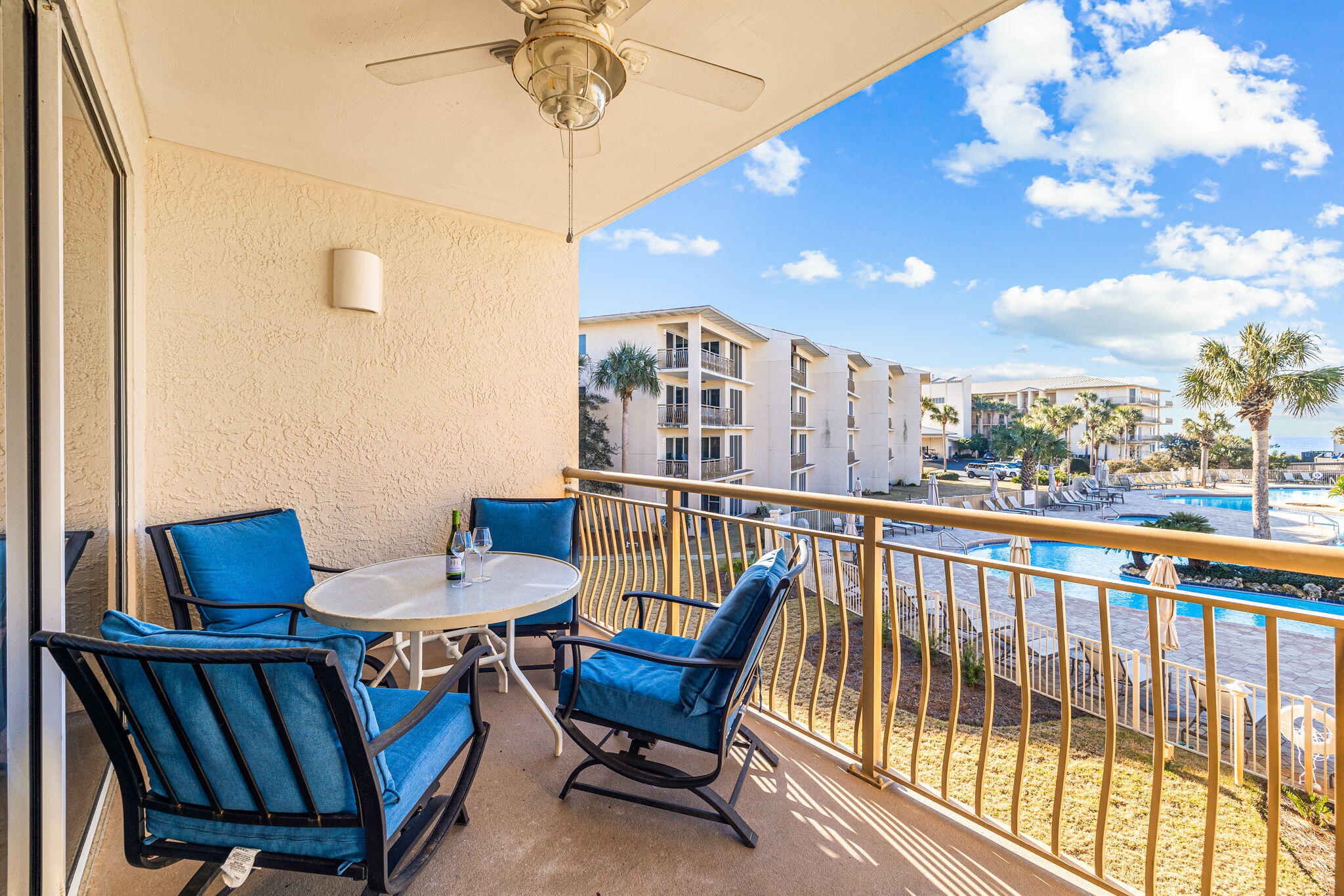 Furnished condo balcony overlooking a pool and palm-lined condominium grounds, with a glimpse of water.
