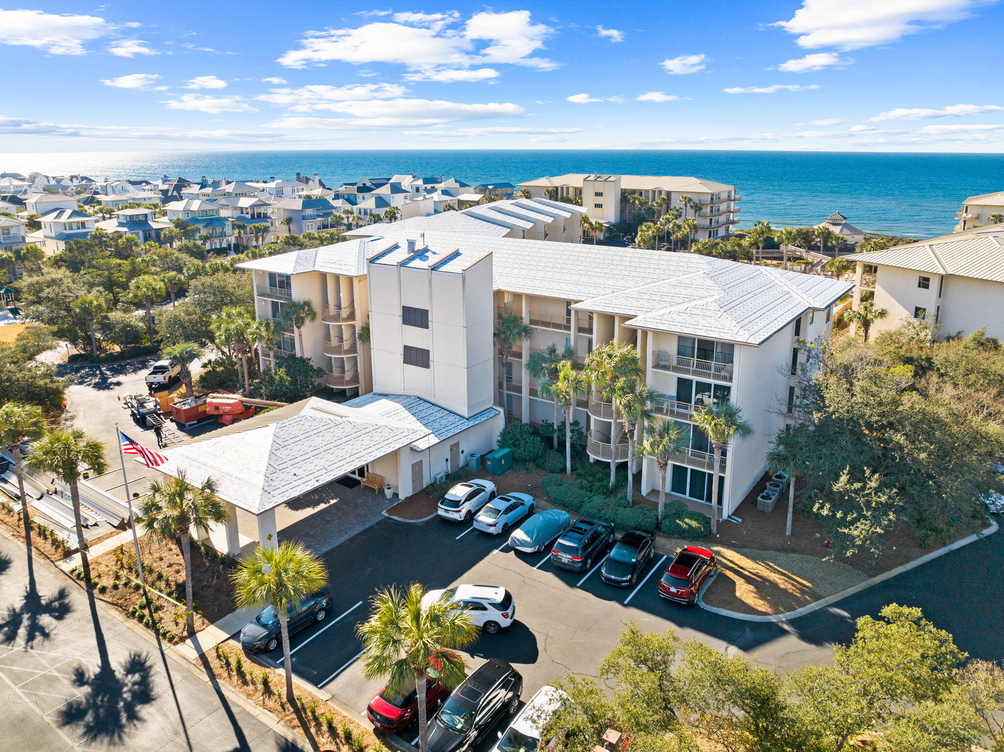 Aerial view of a coastal condominium with palm trees, parking, and the Gulf beyond.