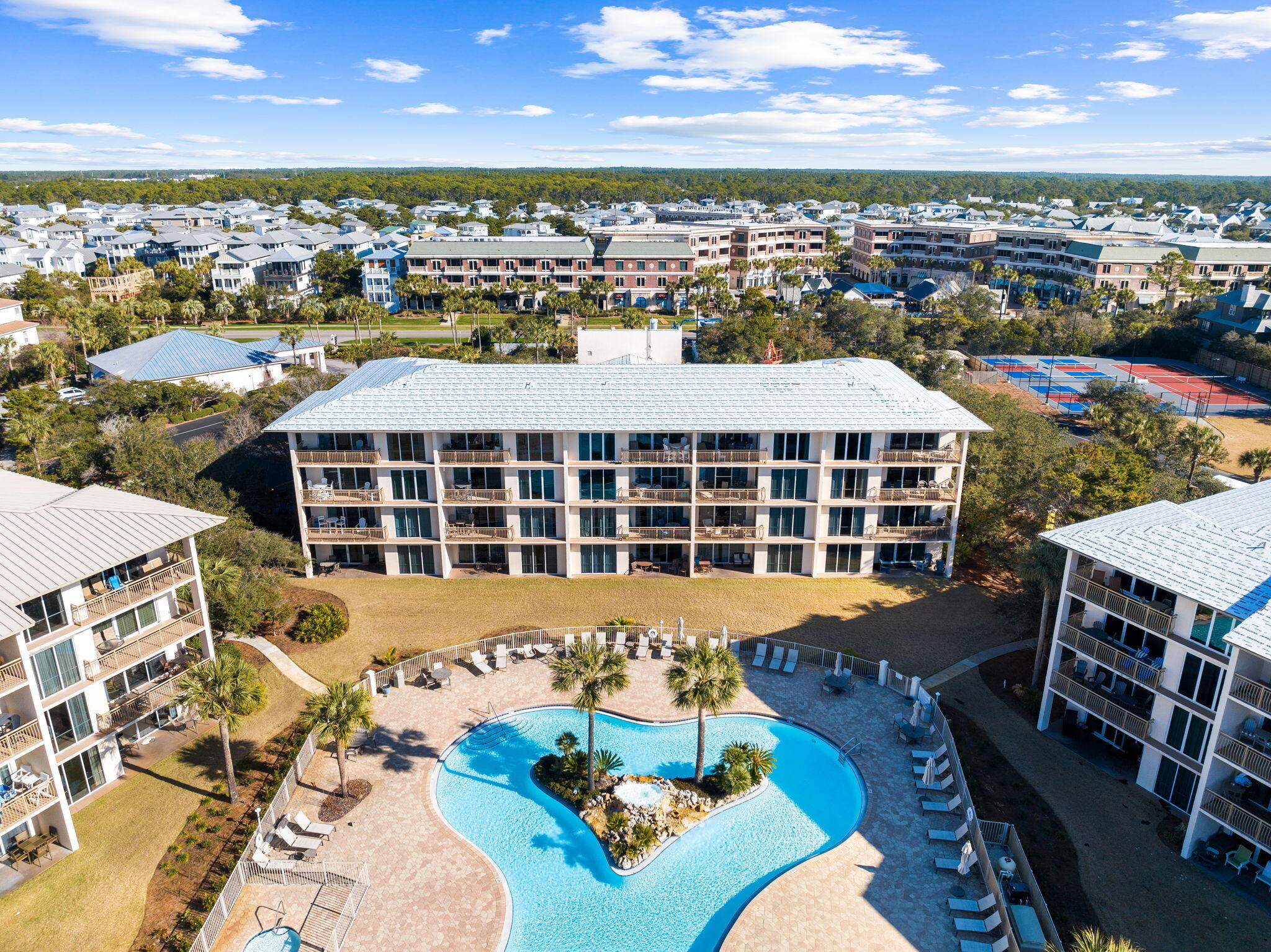 Aerial view of condominium buildings surrounding a landscaped pool courtyard, with tennis courts nearby.