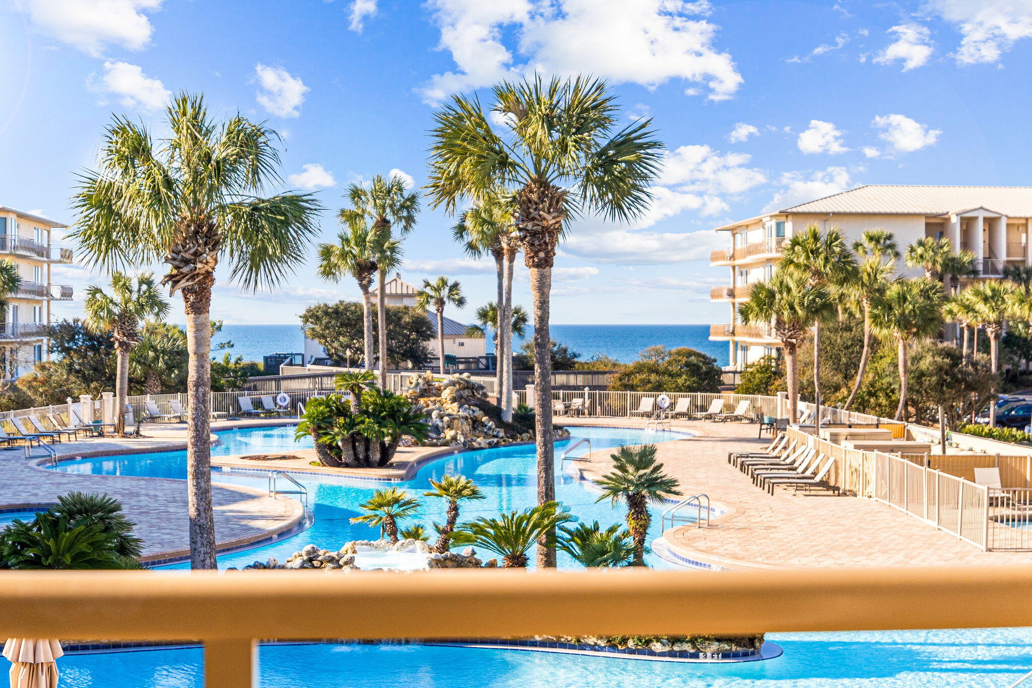 Resort pool with palm trees and lounge chairs, with the ocean visible between condominium buildings.