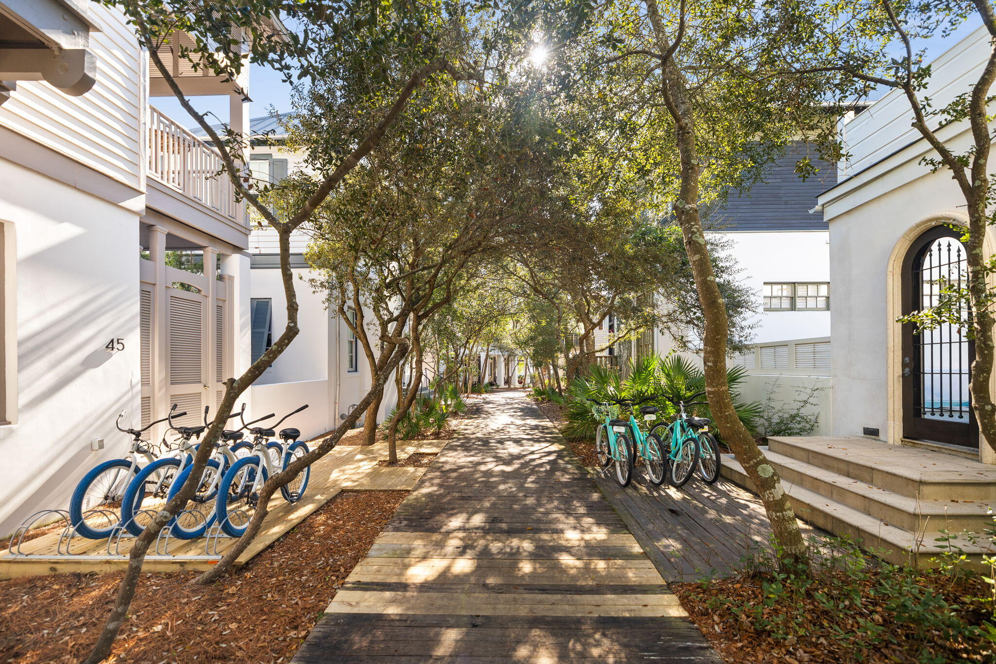 Wooden walkway between white residences, shaded by trees, with bicycles parked along the path.