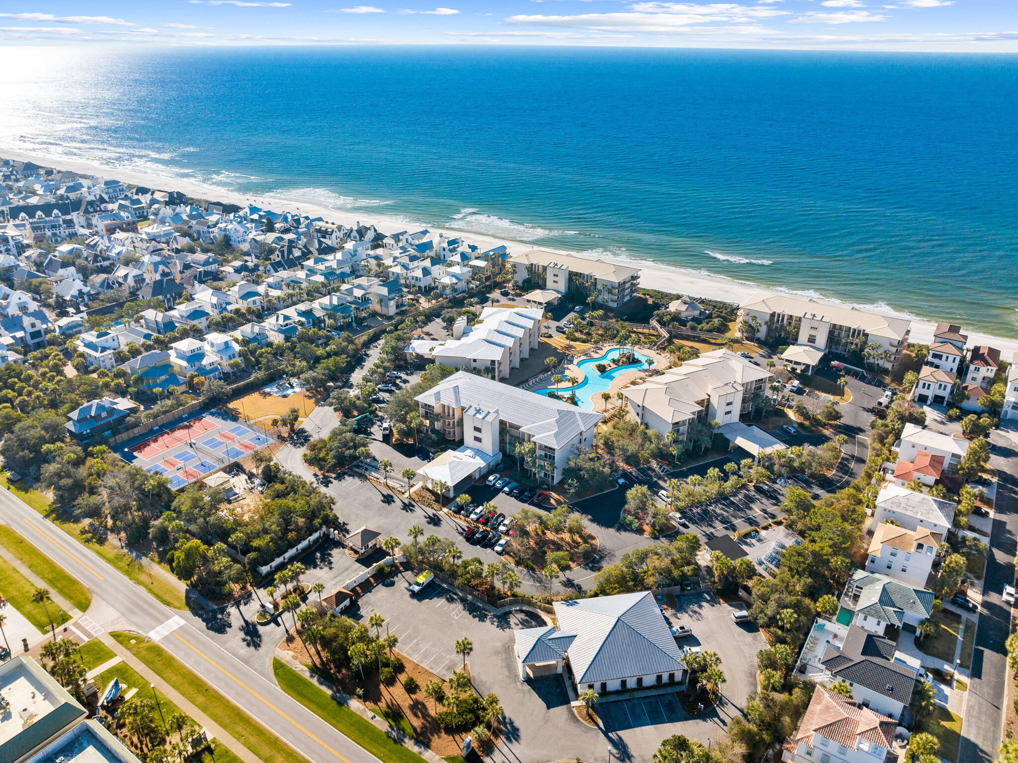 Aerial view of beachfront condominium complex with a pool, tennis courts, and shoreline.