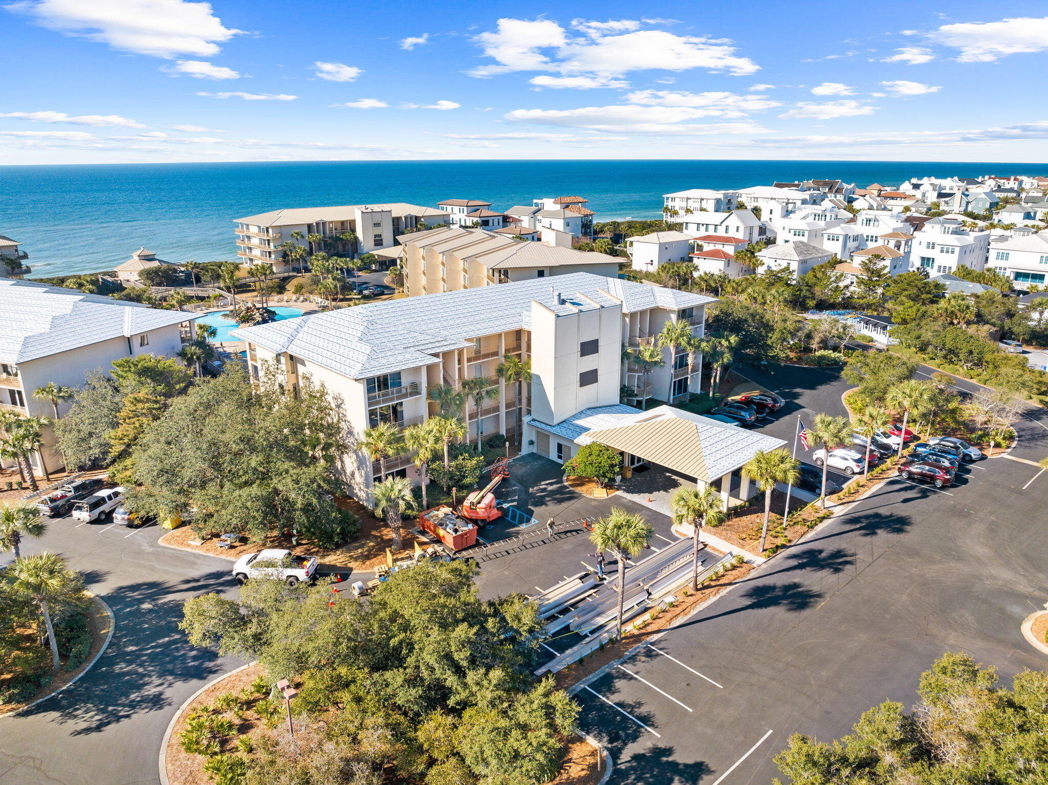 Aerial view of a coastal condominium complex beside the ocean, with a pool and parking.