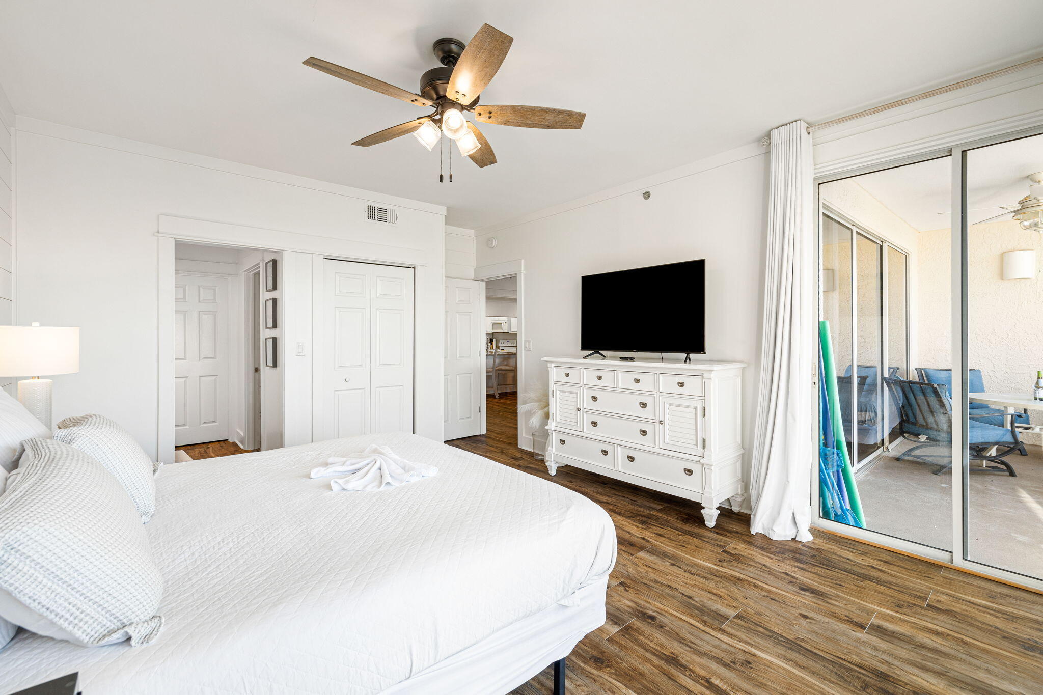 Bedroom with a white bed and dresser beside sliding doors to a furnished balcony.