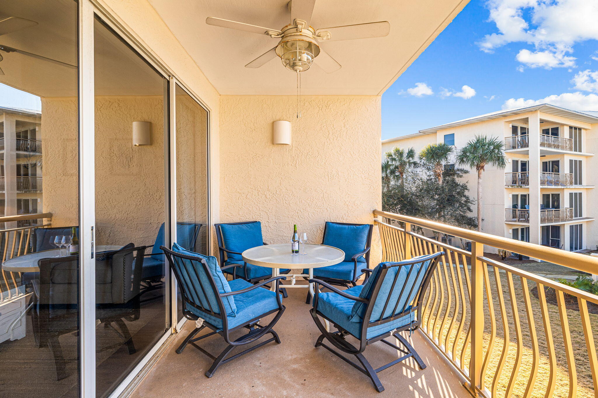 Condo balcony furnished with blue chairs around a round table, overlooking a neighboring building and palm trees.