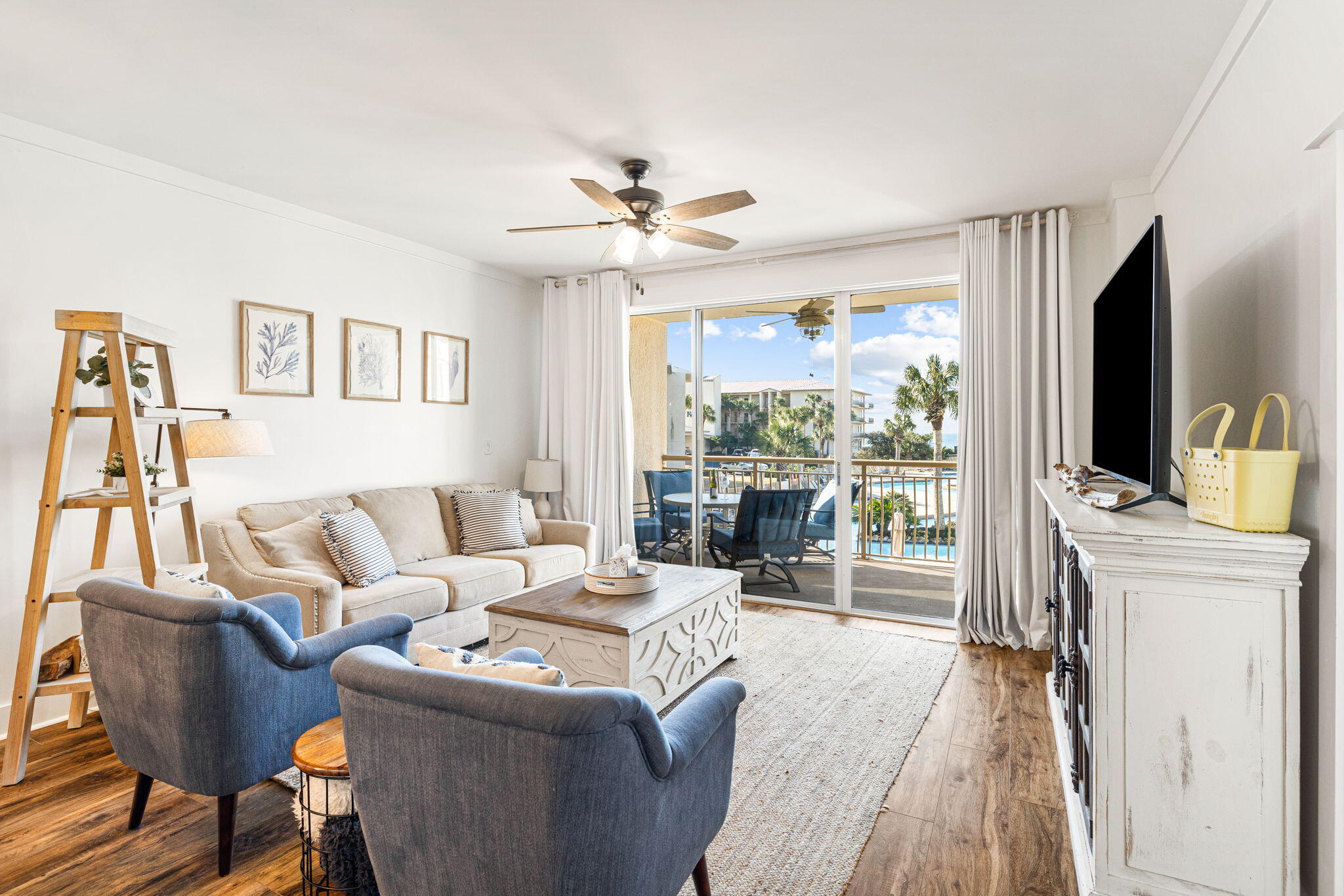 Living room with cream sofa and blue chairs opening onto a balcony with palm trees and water views.