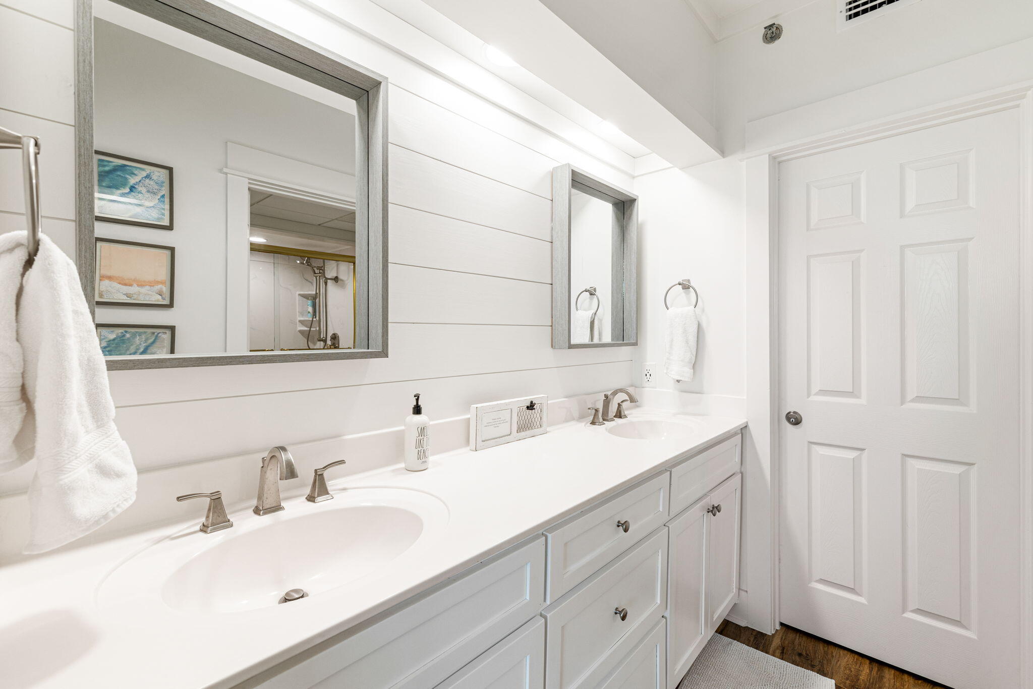 Bright bathroom with a long double-sink vanity, gray-framed mirrors, white shiplap walls, and dark wood flooring.