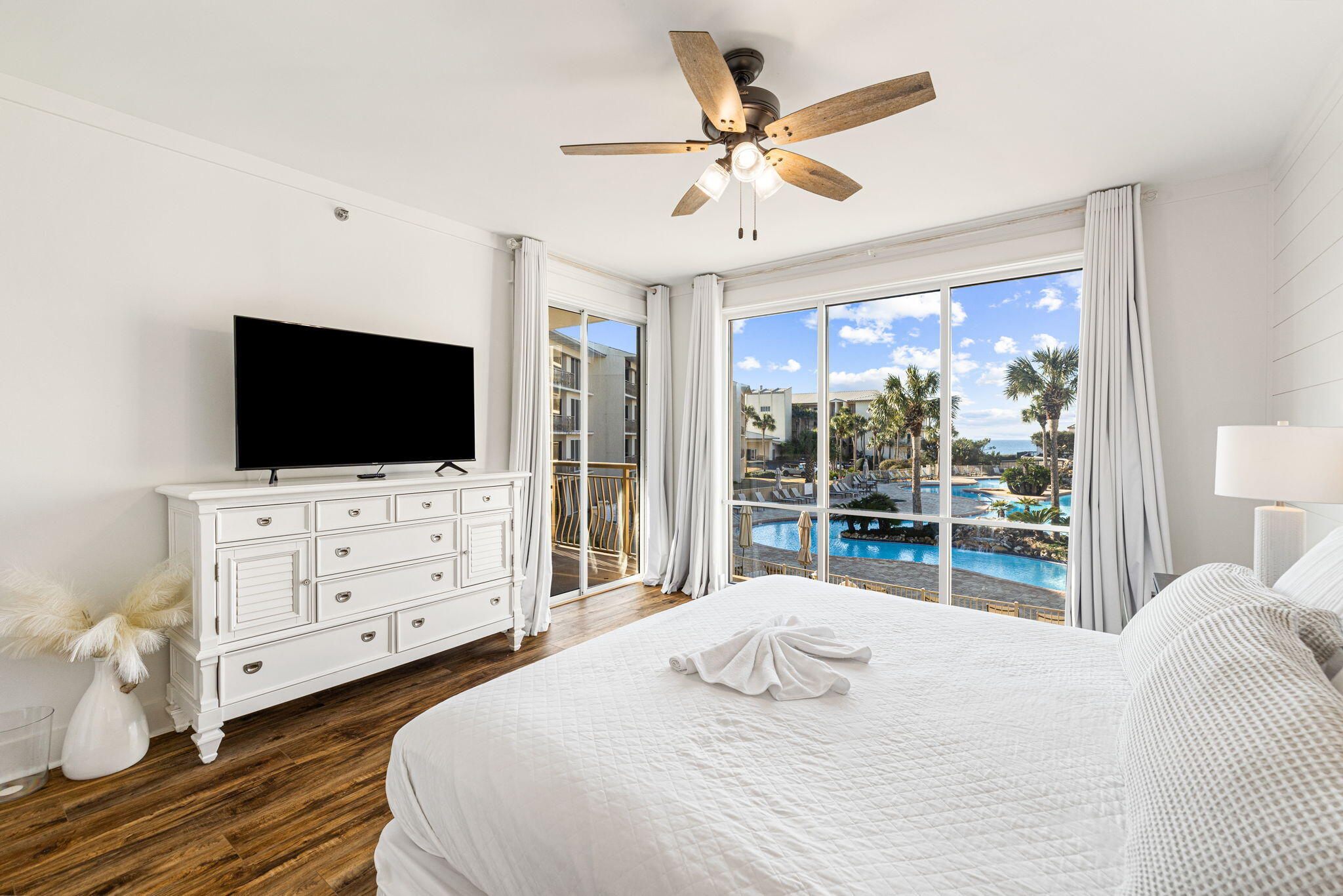 Bedroom with white bedding and dresser, overlooking a palm-lined resort pool through large windows.