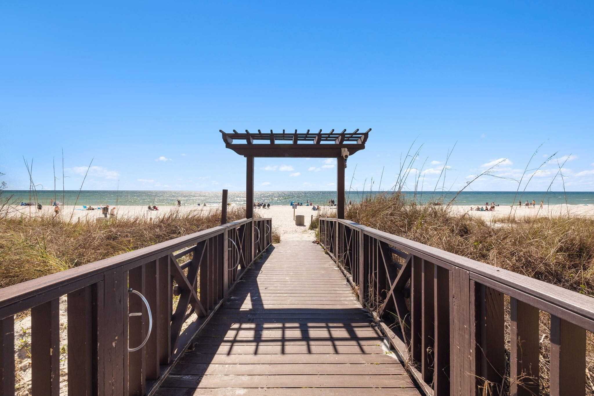 Beach access boardwalk crosses dune grass toward a sunny sandy beach and ocean.