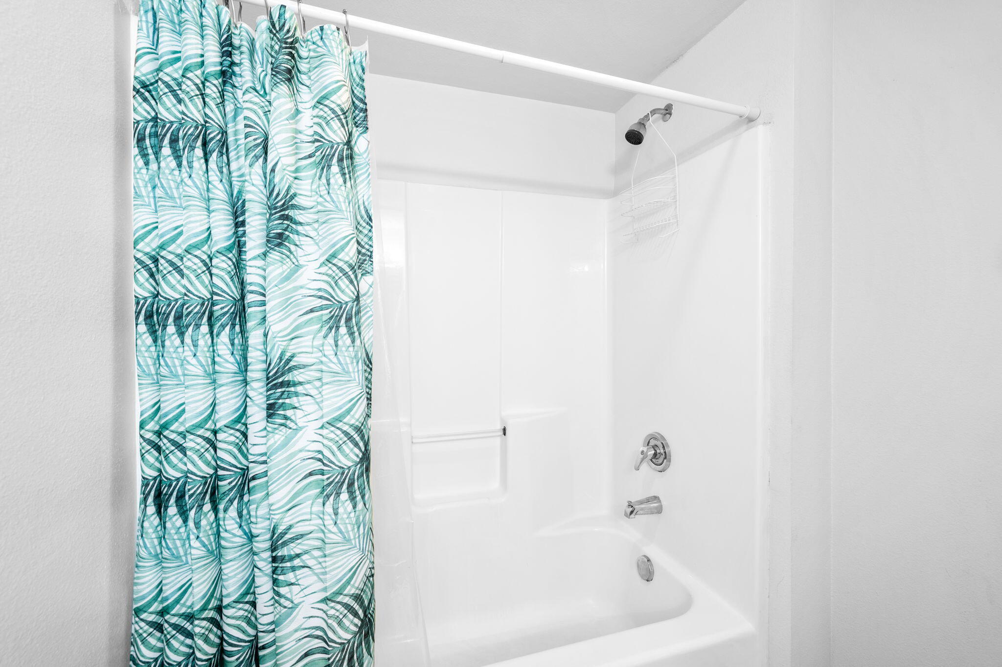 White bathtub and shower with chrome fixtures and a teal tropical-leaf curtain.
