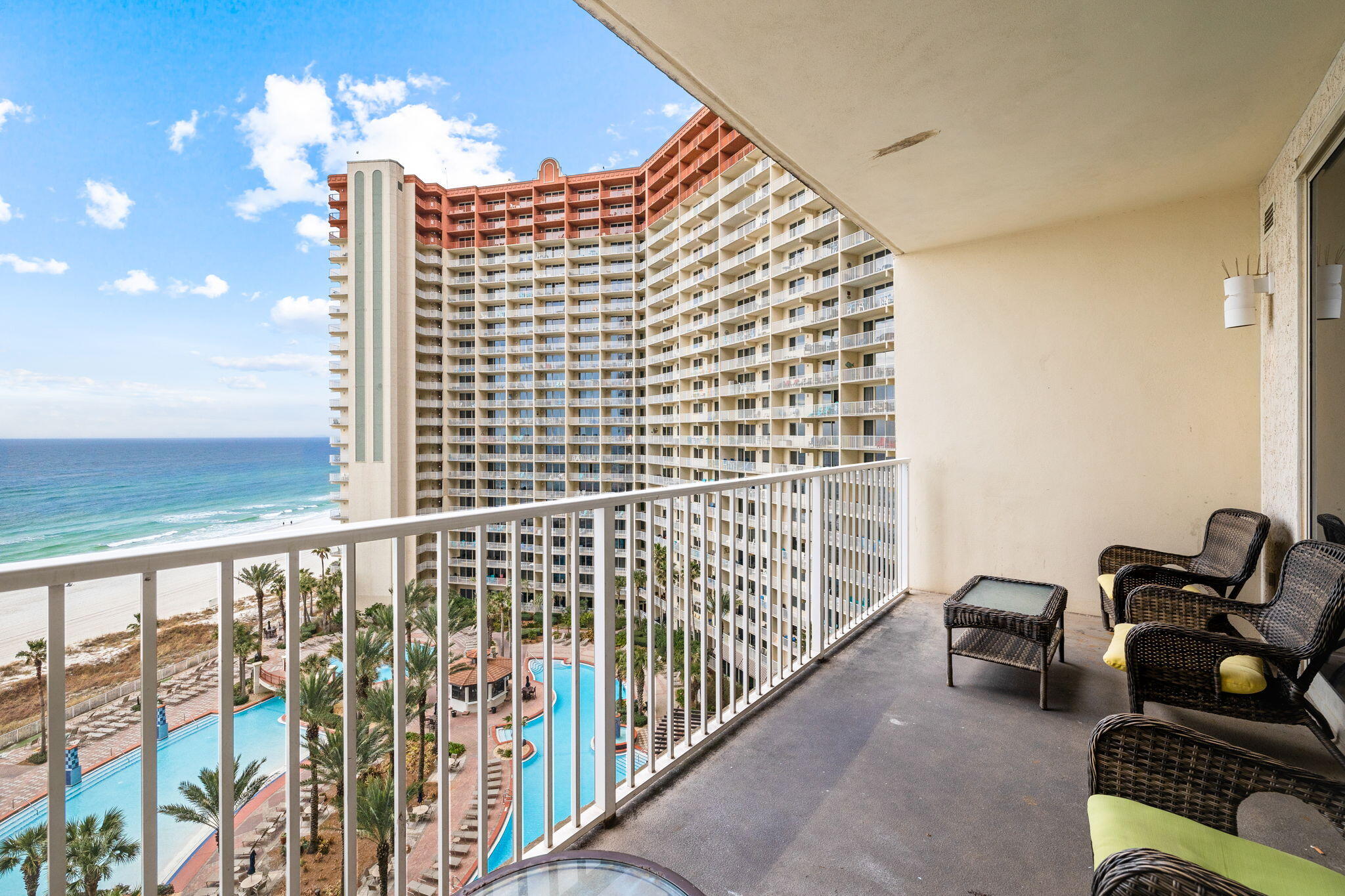 Condo balcony with wicker seating overlooking a pool, beach, ocean, and neighboring high-rise.