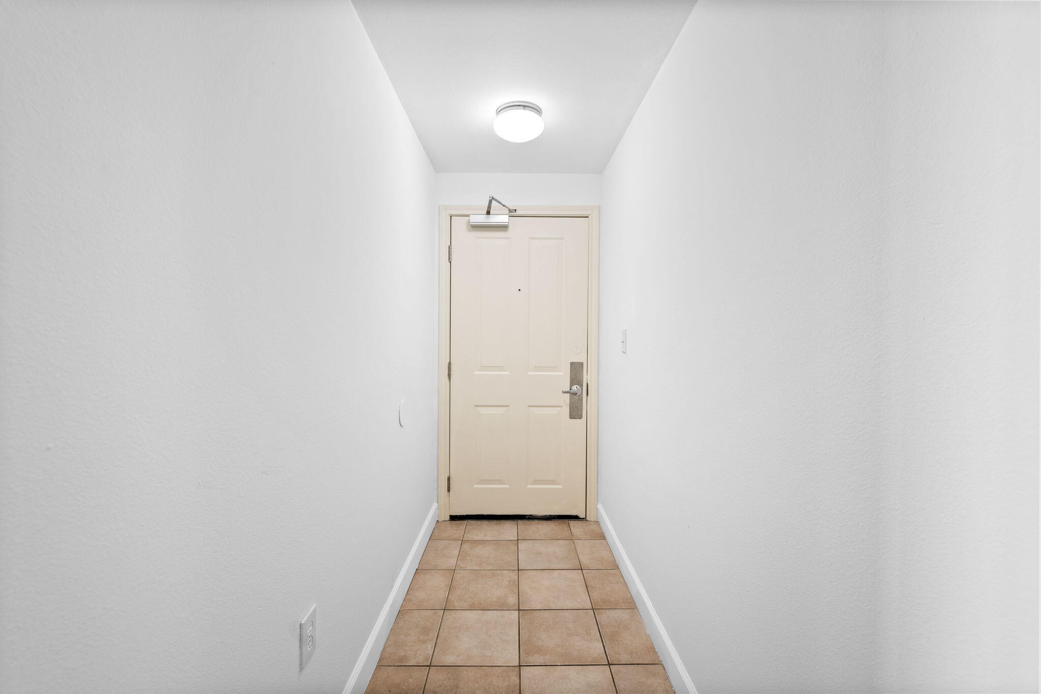 Empty condo entry hallway with tan tile flooring and a beige front door.
