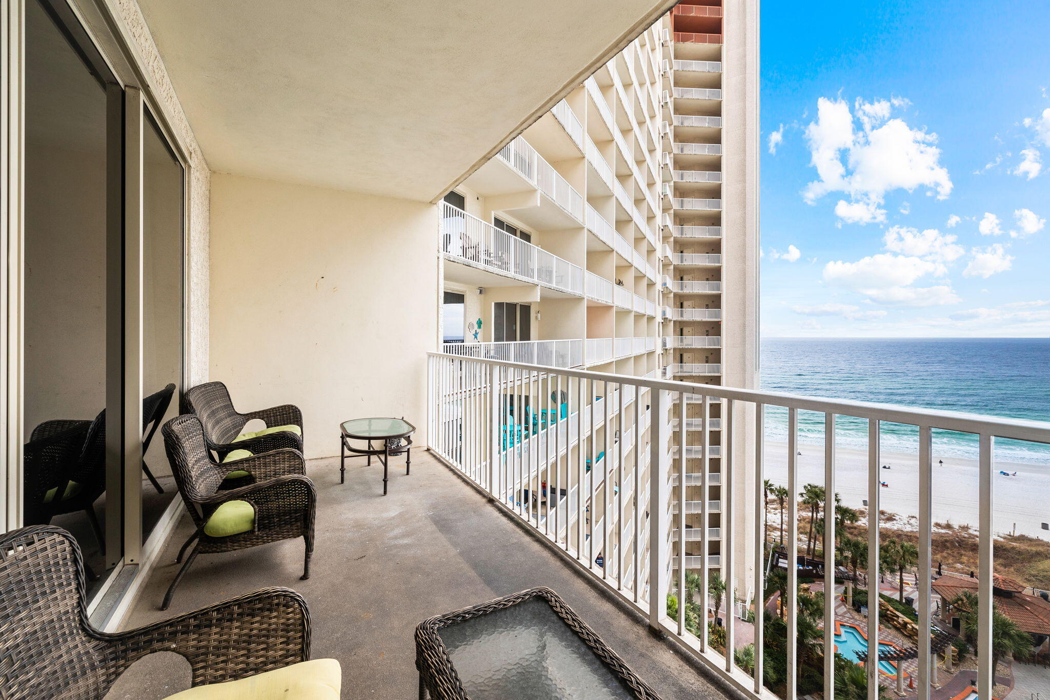 Furnished condo balcony overlooking the beach and ocean, with wicker chairs and neighboring balconies.