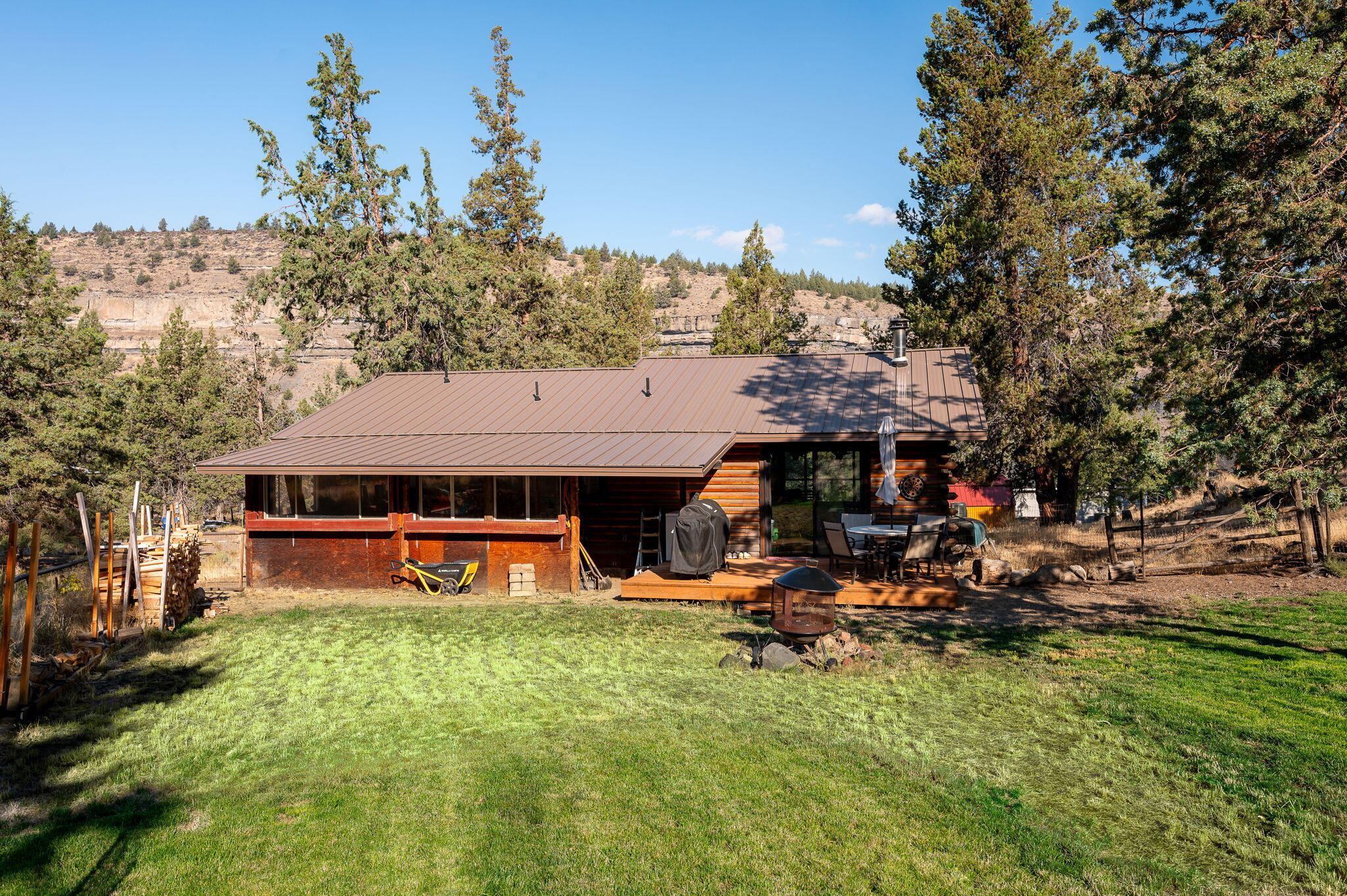 Rear of a log-sided home with a deck, lawn, mature trees, and rocky canyon cliffs.
