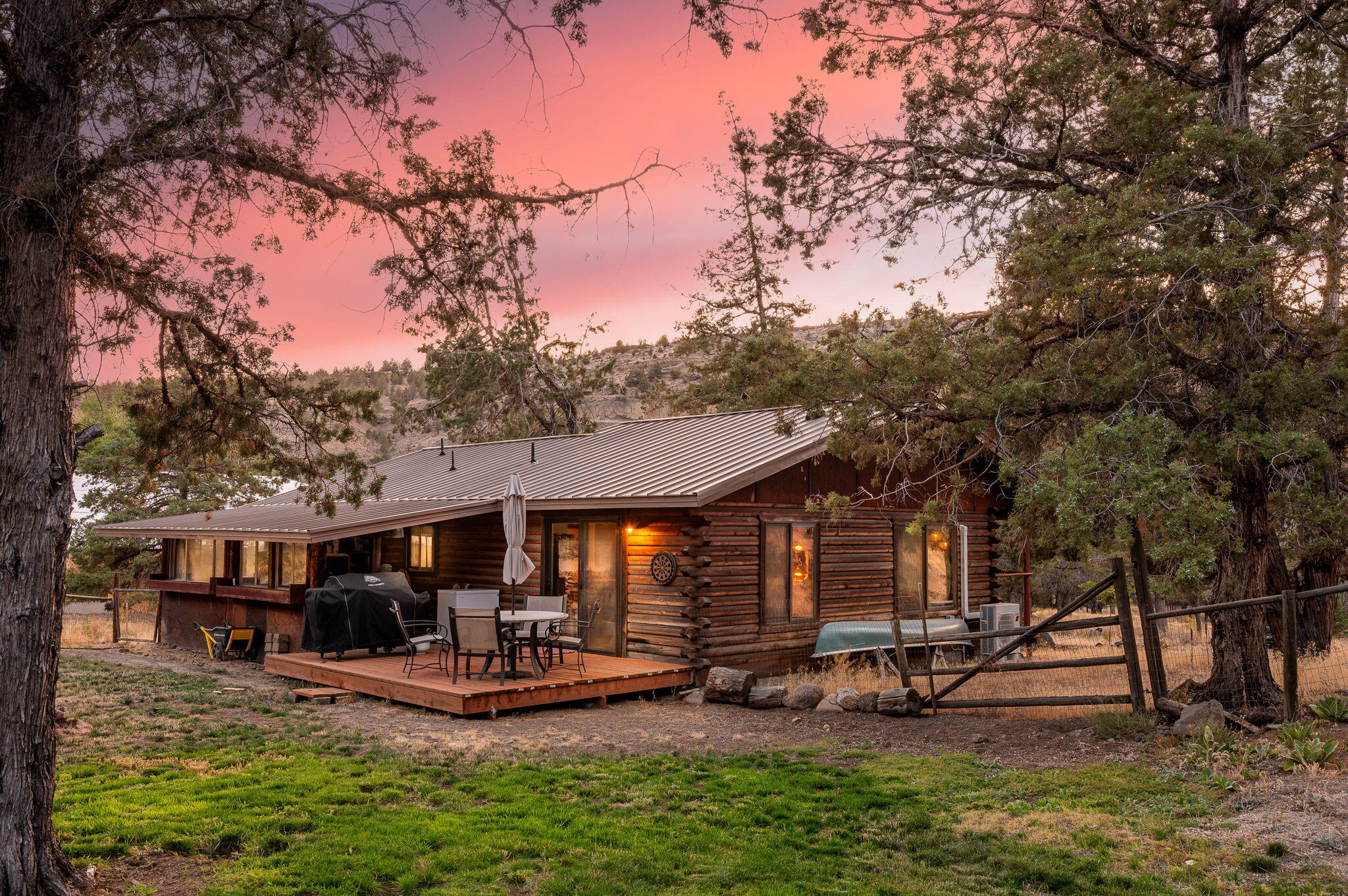 Log cabin with a metal roof and furnished deck framed by trees at dusk.