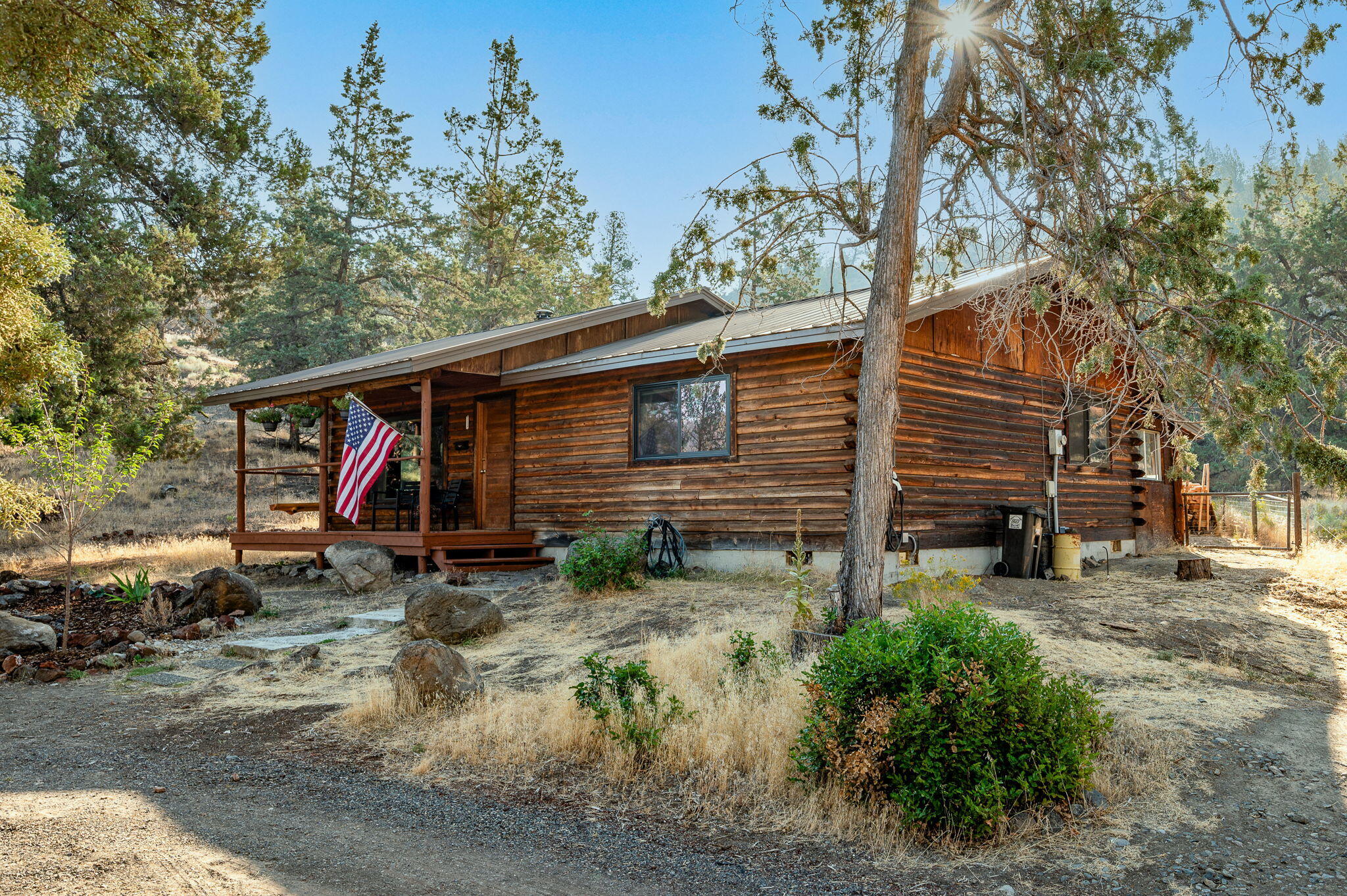 Rustic log cabin with a covered porch and American flag, surrounded by trees and dry grass.