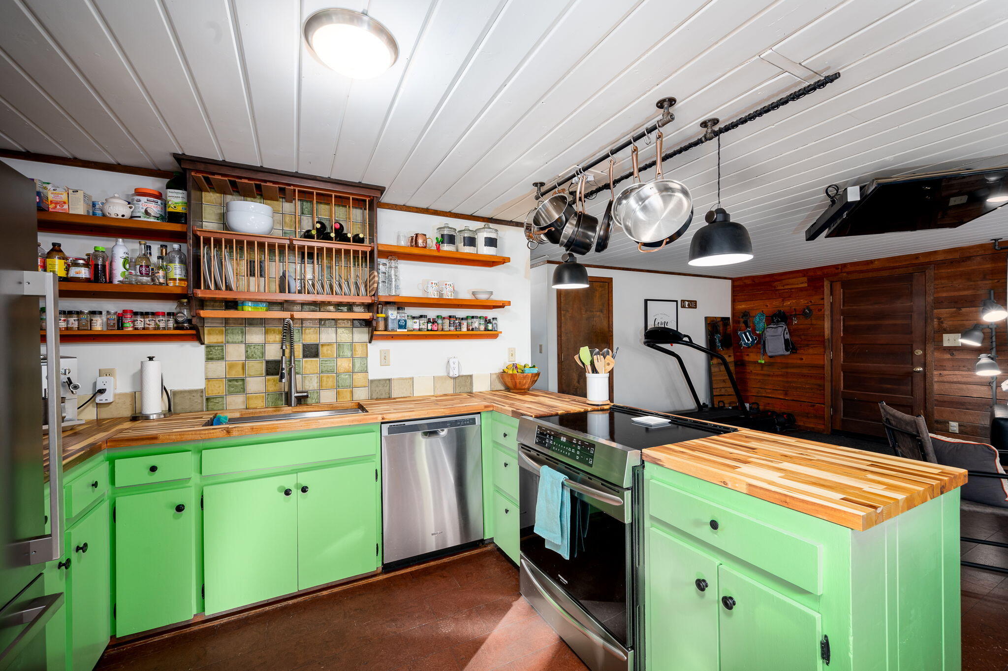 Kitchen with bright green cabinets, butcher-block counters, and hanging pots opening into a wood-paneled room.