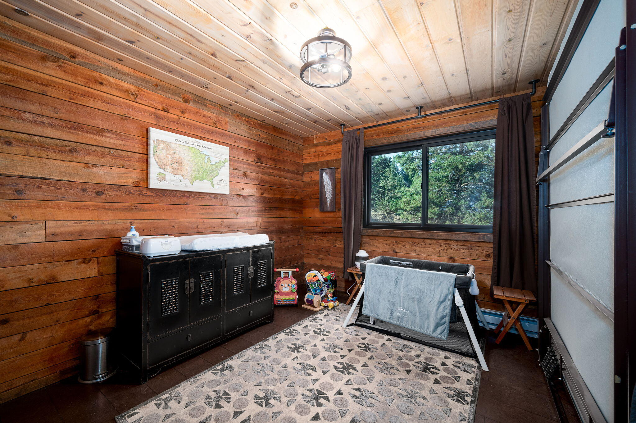 Wood-paneled nursery with a changing cabinet, baby playpen, toys, and a window overlooking trees.