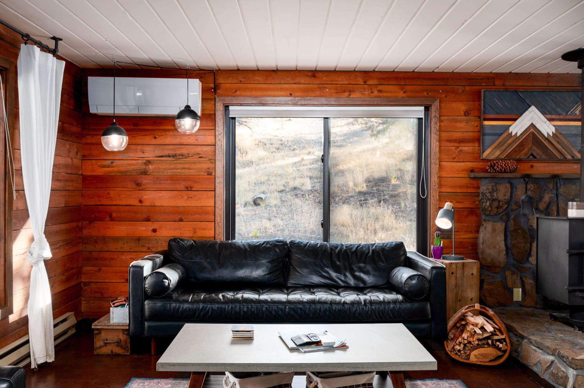 Living room with wood walls, a black leather sofa, and a stone fireplace beside a large window.