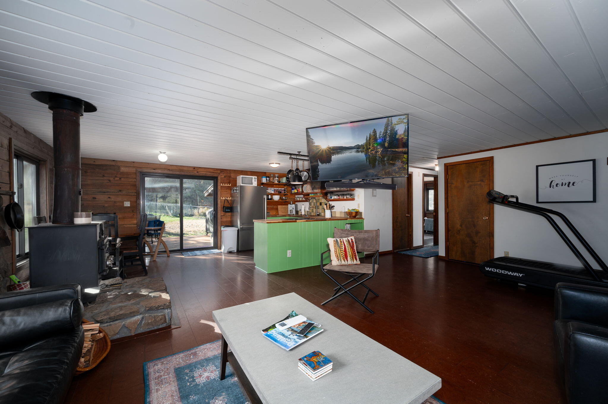 Open-plan kitchen and living room with a green island, wood-paneled walls, and a black wood stove.