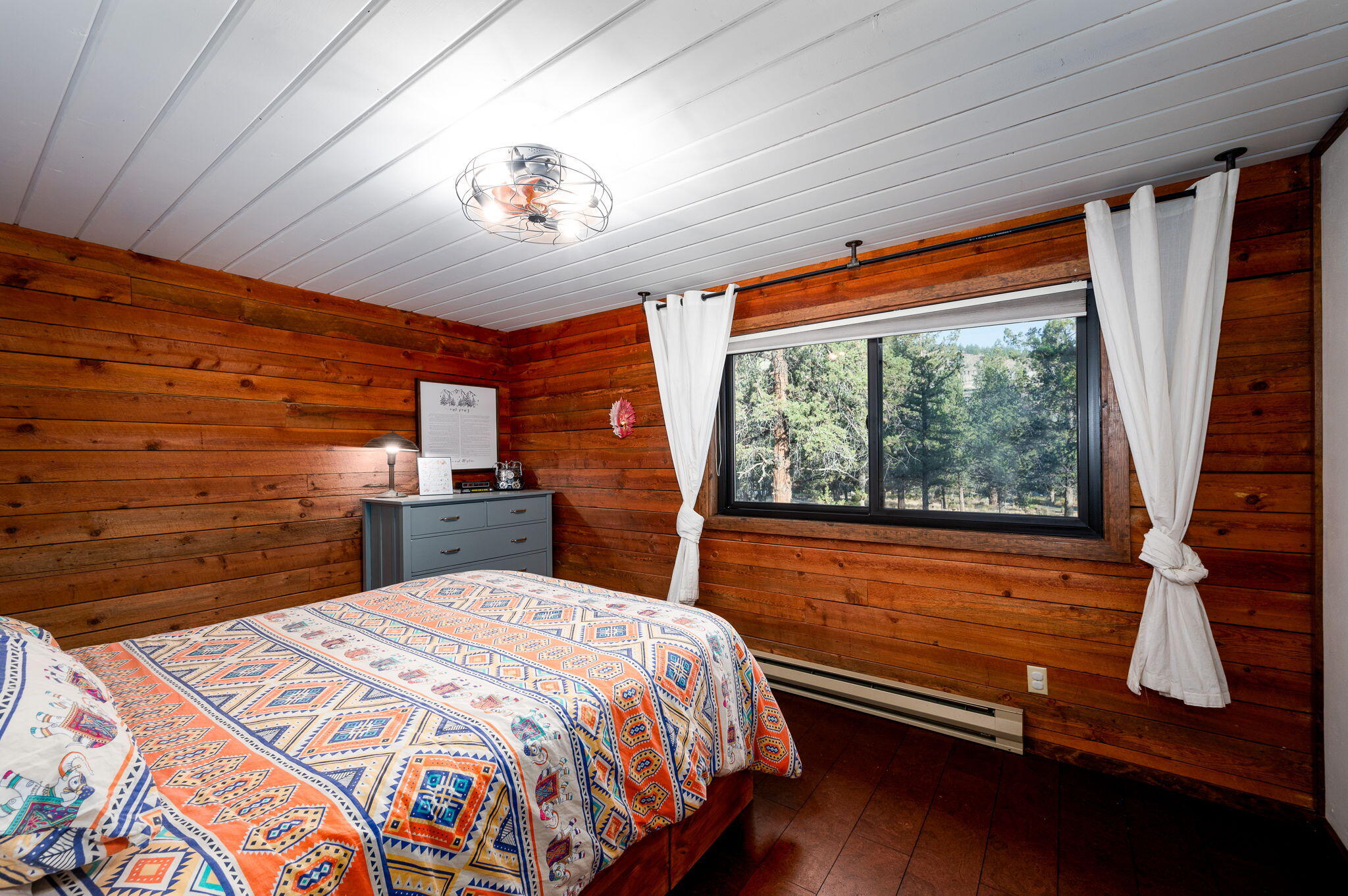 Bedroom with wood-plank walls, a colorful geometric bedspread, and a window overlooking trees.