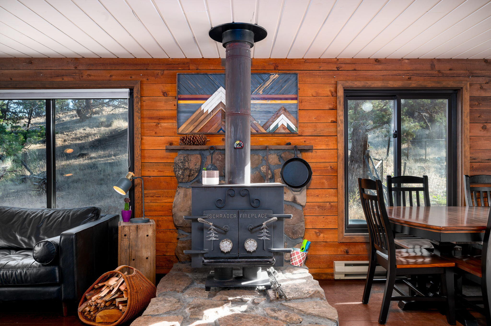 Wood-burning fireplace between a black leather sofa and dining table, with wood walls and windows overlooking trees.