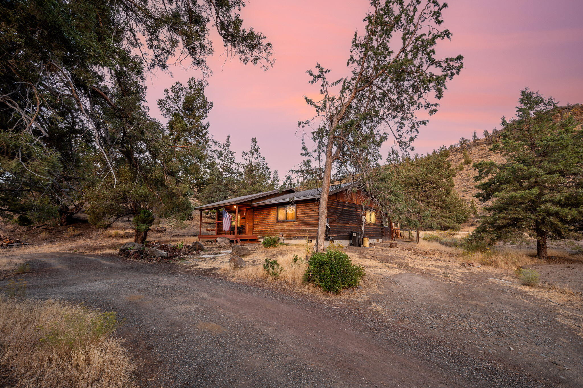 Log cabin with a covered porch and glowing windows among trees beneath a pink dusk sky.