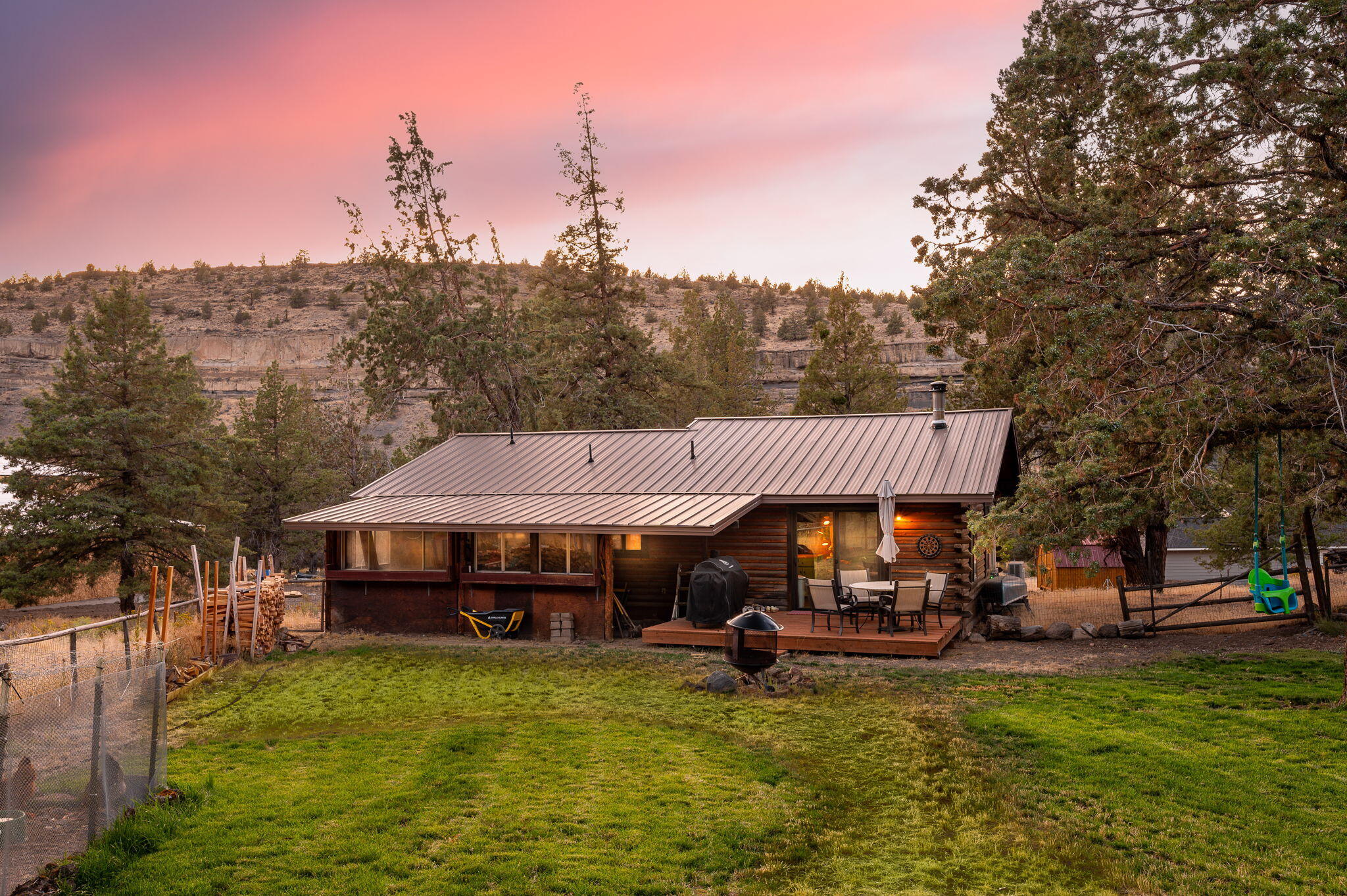 Rustic log-sided home with a metal roof and furnished deck beneath rocky canyon cliffs at sunset.