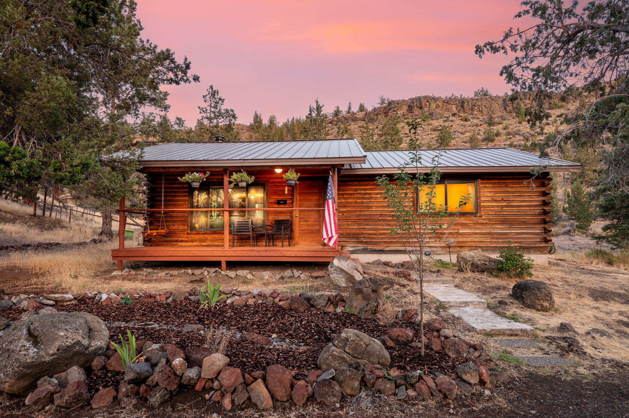 Log cabin with a metal roof and front porch, framed by rocky landscaping beneath a pink dusk sky.