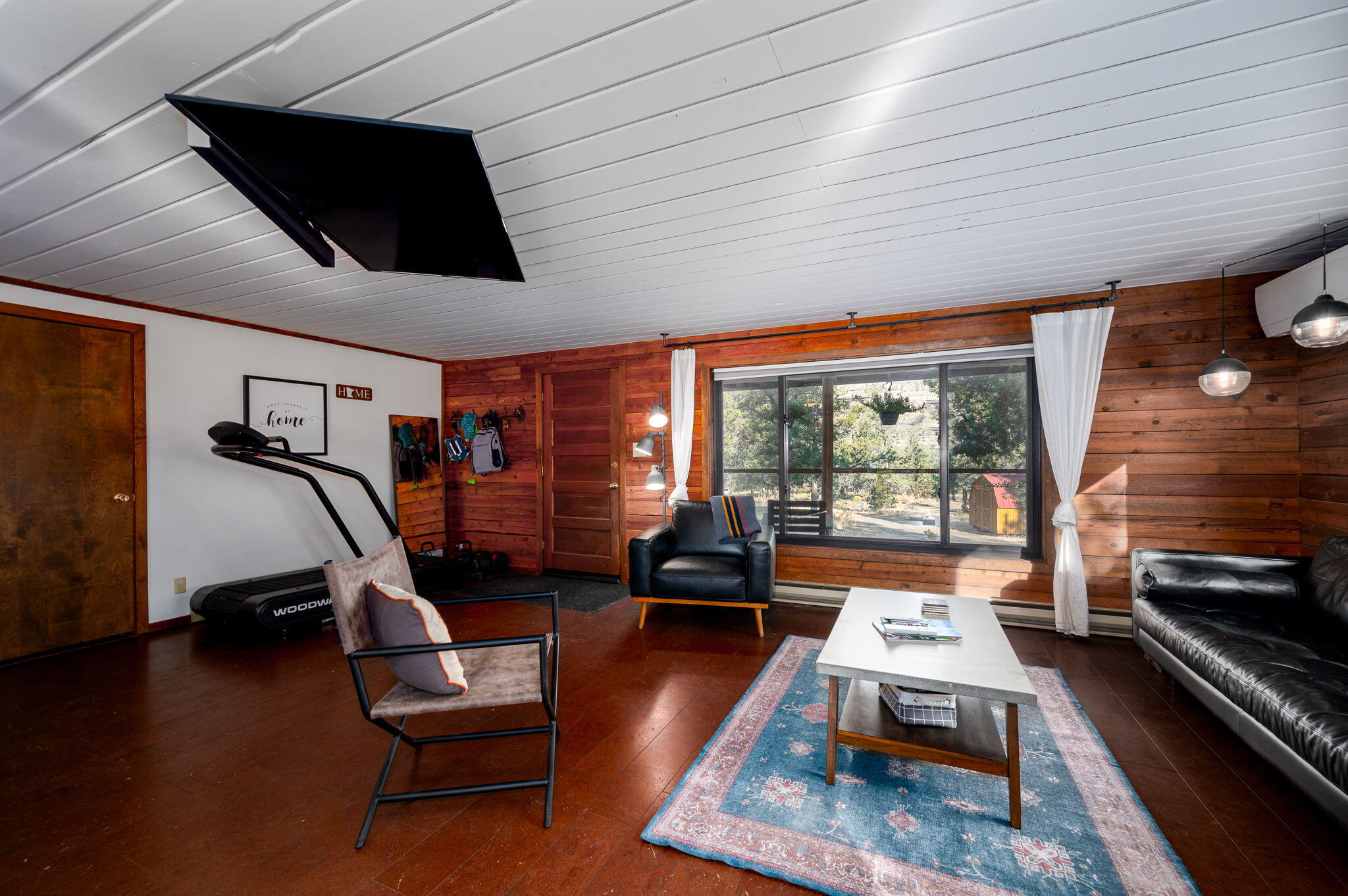 Living room with wood-paneled walls, a large curtained window, seating, and a treadmill.