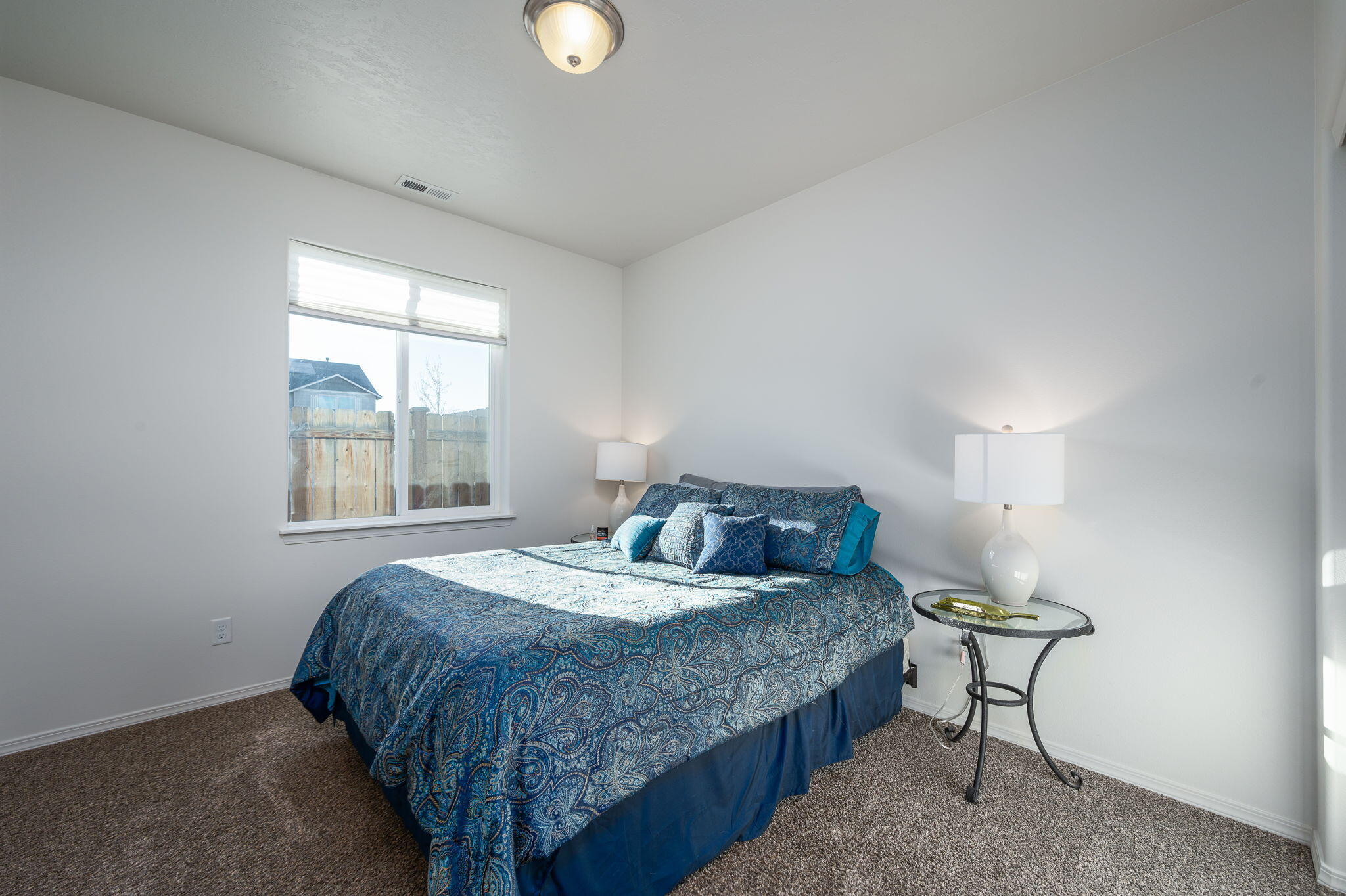 Bedroom with blue paisley bedding, gray carpet, and a window overlooking a fenced yard.