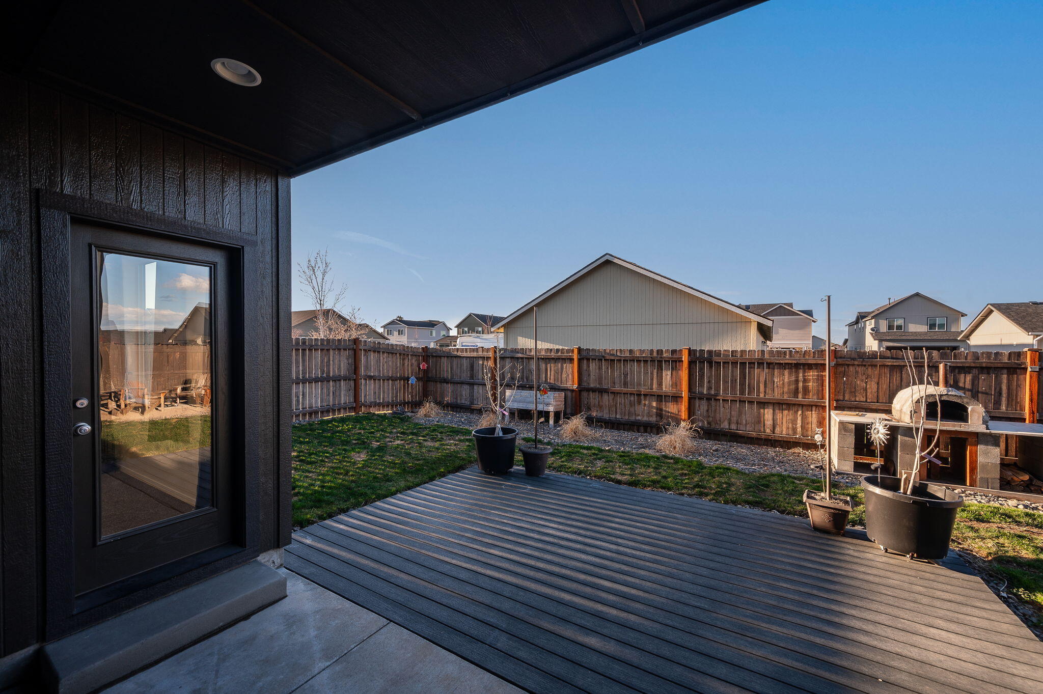 Covered backyard patio with a dark deck, fenced lawn, potted plants, and an outdoor oven.